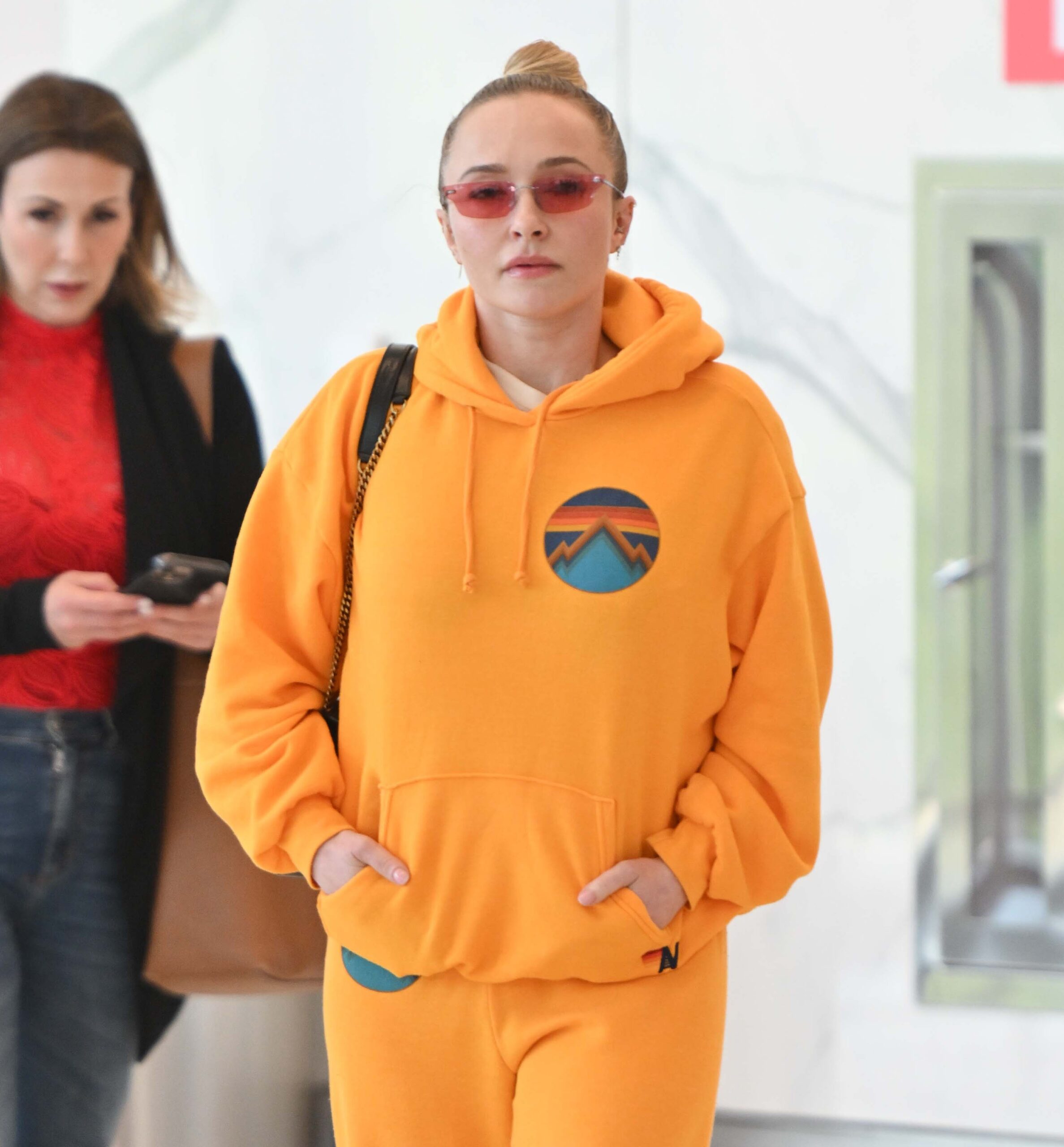 Hayden Panettiere in an orange Aviator Nation hoodie and sweatpants walking through LaGuardia Airport alongside her publicist