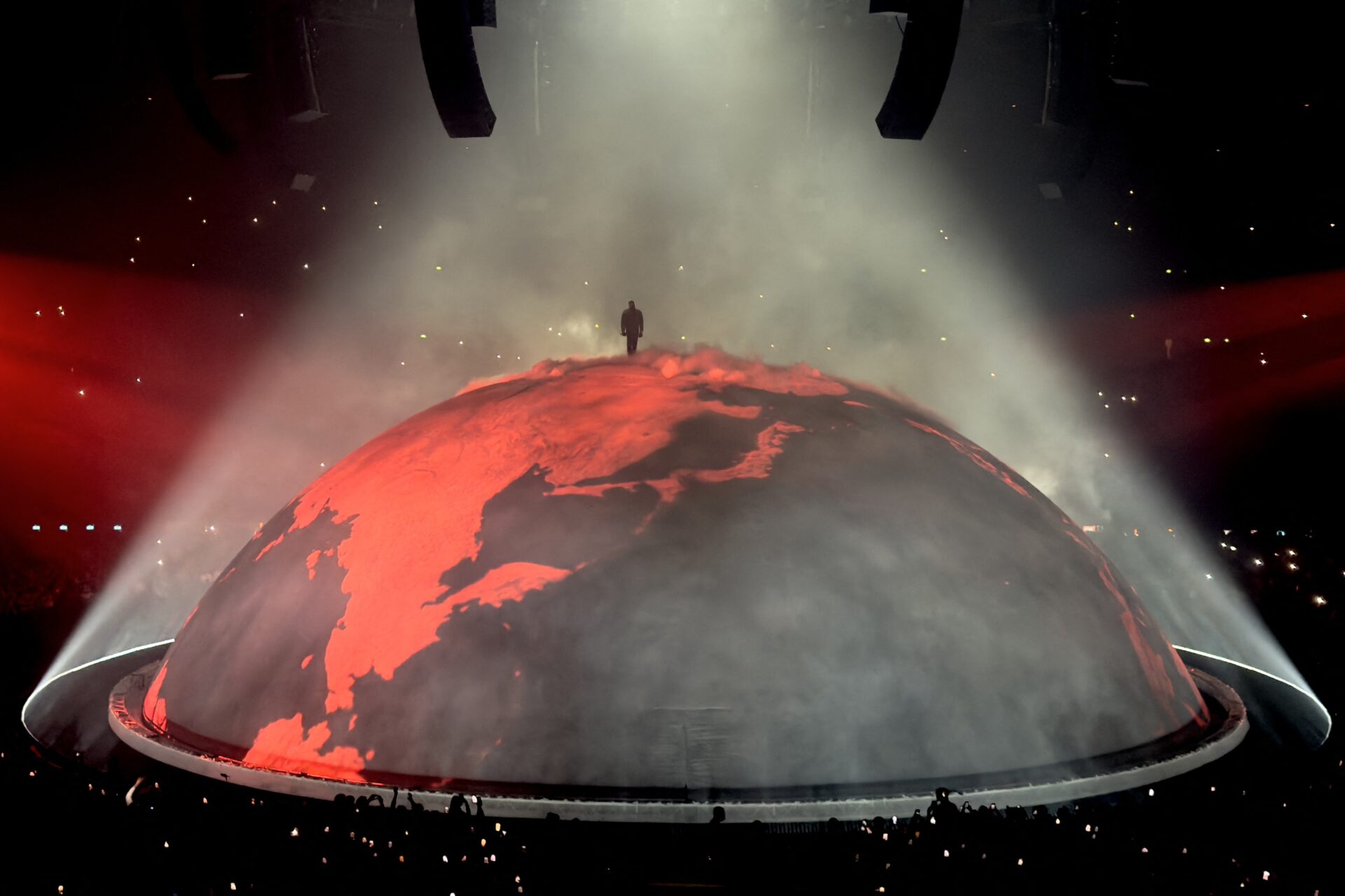 Silhouette of a performer standing on top of a large red glowing dome-shaped stage set, lit by a spotlight with fog filling a darkened arena.