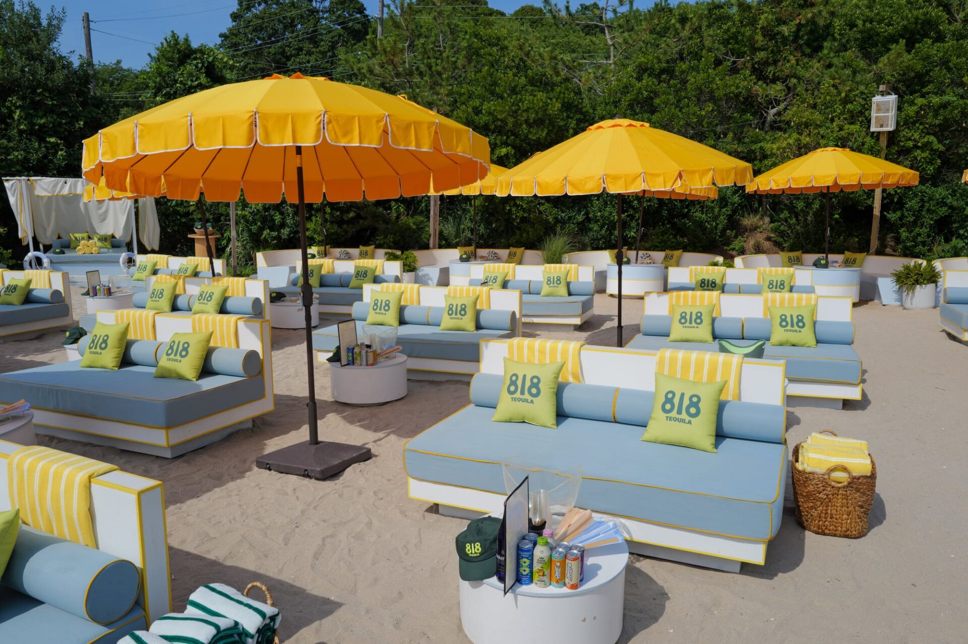 Blue-and-white lounge chairs decorated with 818 Tequila pillows and yellow towels sit beneath bright yellow umbrellas at a sandy beach club.