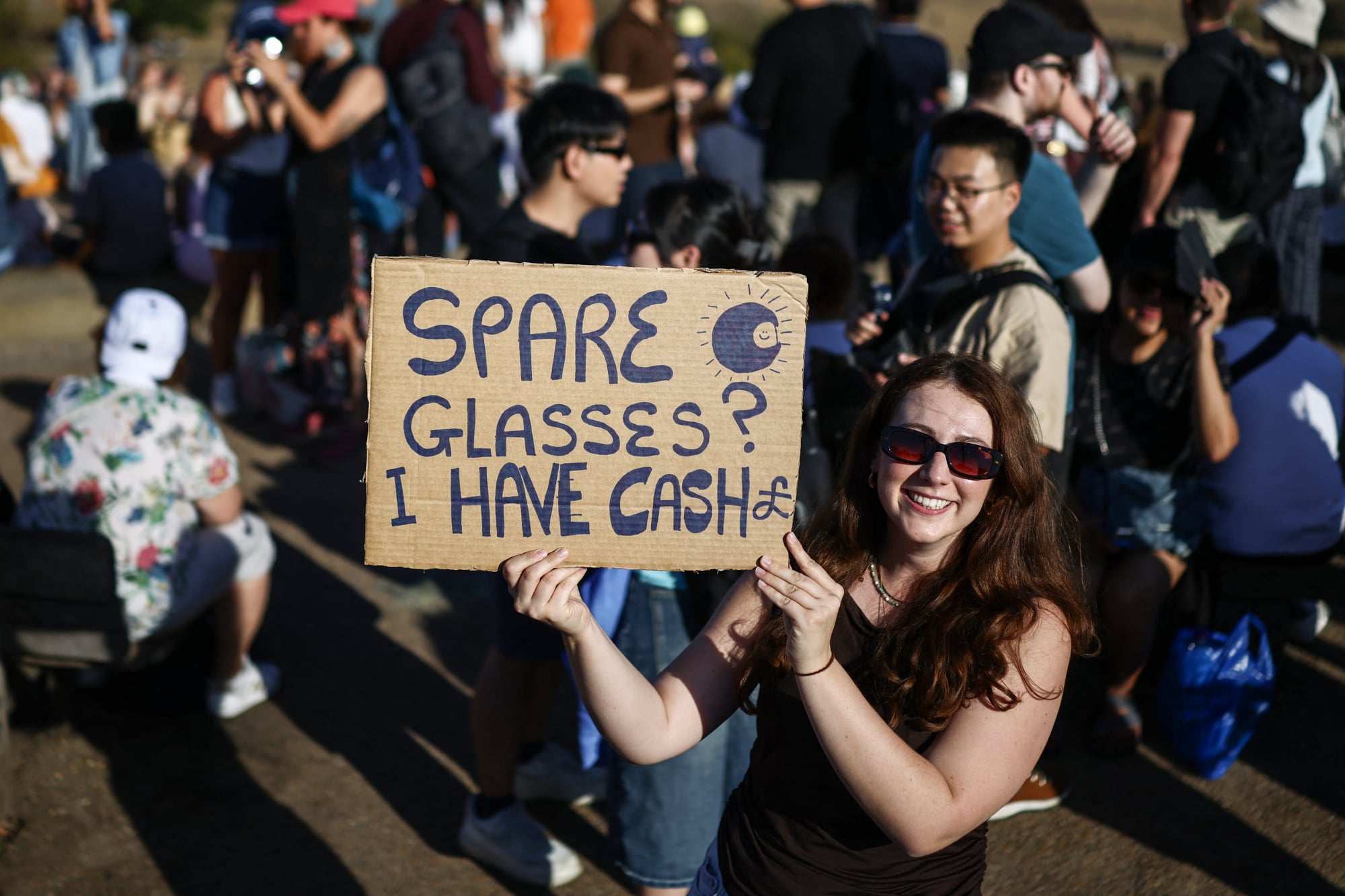 A woman holds a sign in front of crowd. It reads "solar eclipse glasses? I have cash!"
