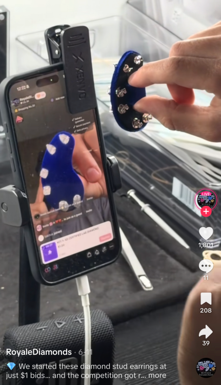 A hand holds diamond stud earrings on a blue velvet display beside a phone streaming a Royale Diamonds TikTok Live auction.