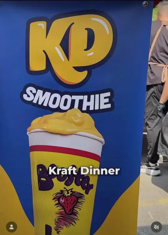 A blue sign advertising a “KD Smoothie” shows an illustrated yellow drink topped with a thick layer of cheese sauce.