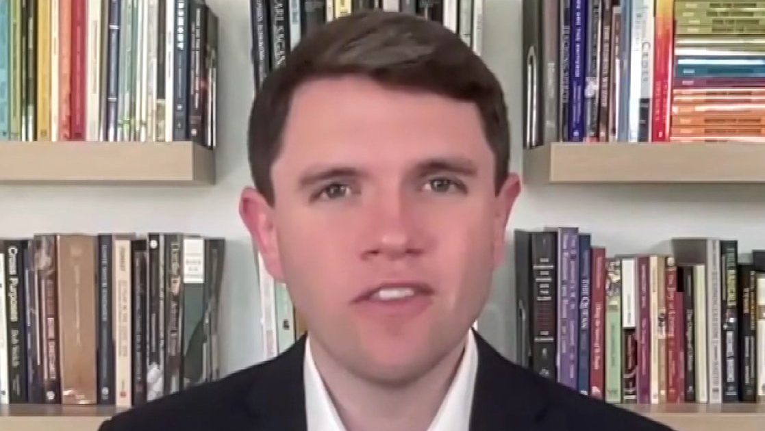 Man in a suit smiling in front of a bookshelf filled with various books, showcasing a professional and academic environment.