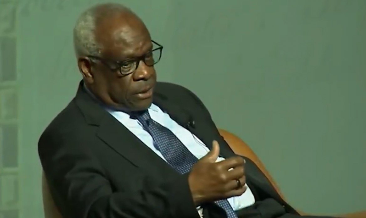 A distinguished speaker in a suit engages in a thoughtful discussion, emphasizing key points with hand gestures during a public event.