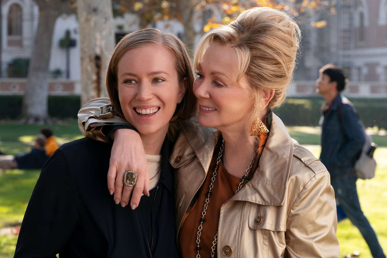 Two women — one younger with brown hair, one older with an upswept blonde hairdo — smile closely together outdoors on a college campus green.