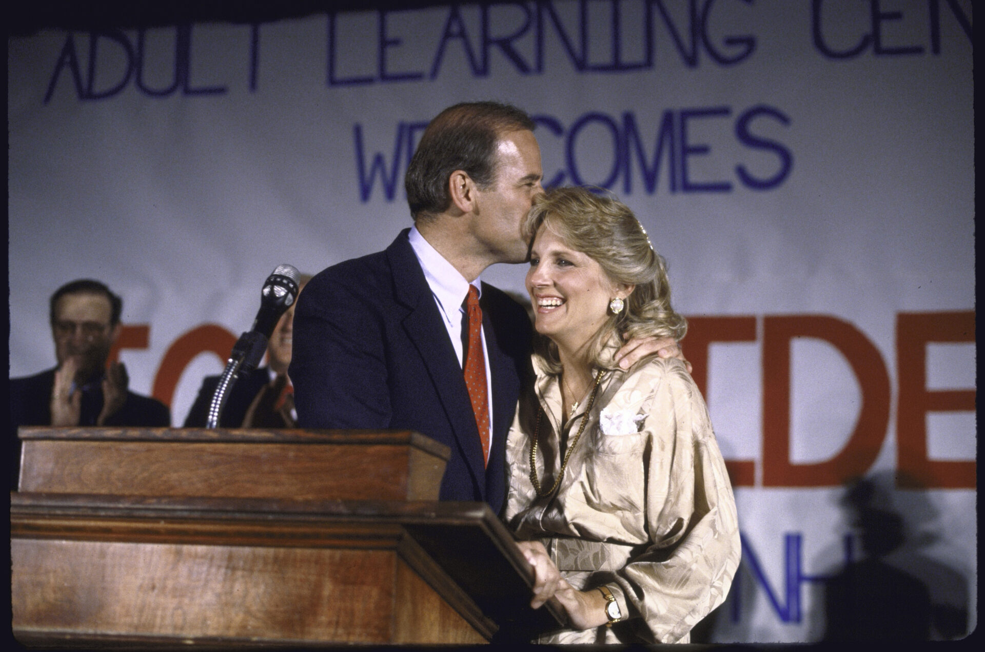 A man in a dark suit kisses a smiling blonde woman on the forehead at a wooden podium, with a welcome banner visible behind them.