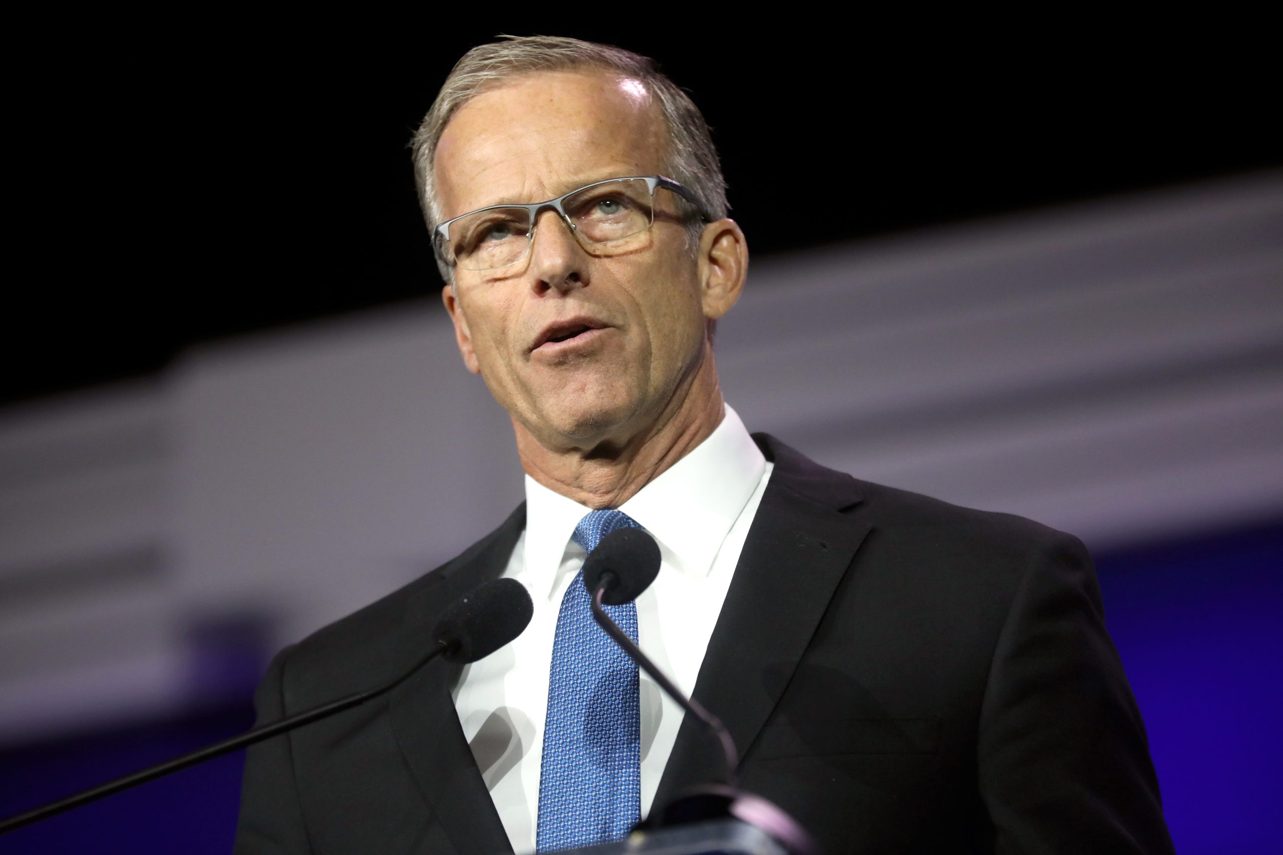 Business professional delivering a speech at a conference, wearing glasses and a suit, with microphones in front of him.
