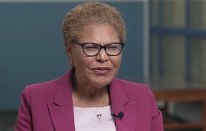 Woman with short curly hair wearing glasses and a pink blazer, speaking in a professional setting.