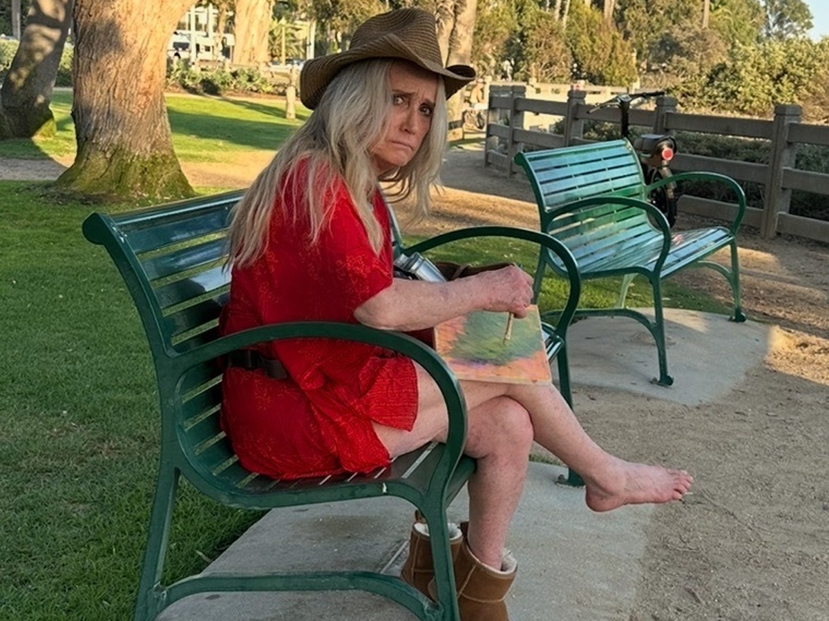 Kim Richards sitting on a park bench near Santa Monica Pier, wearing a cowboy hat and red dress with one boot on and one foot bare, painting