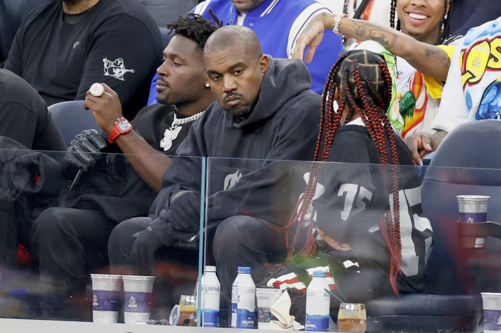 Kanye West seated between Antonio Brown and a person with red braids at Super Bowl LVI, with cups and Aquafina bottles on the ledge in front of them.