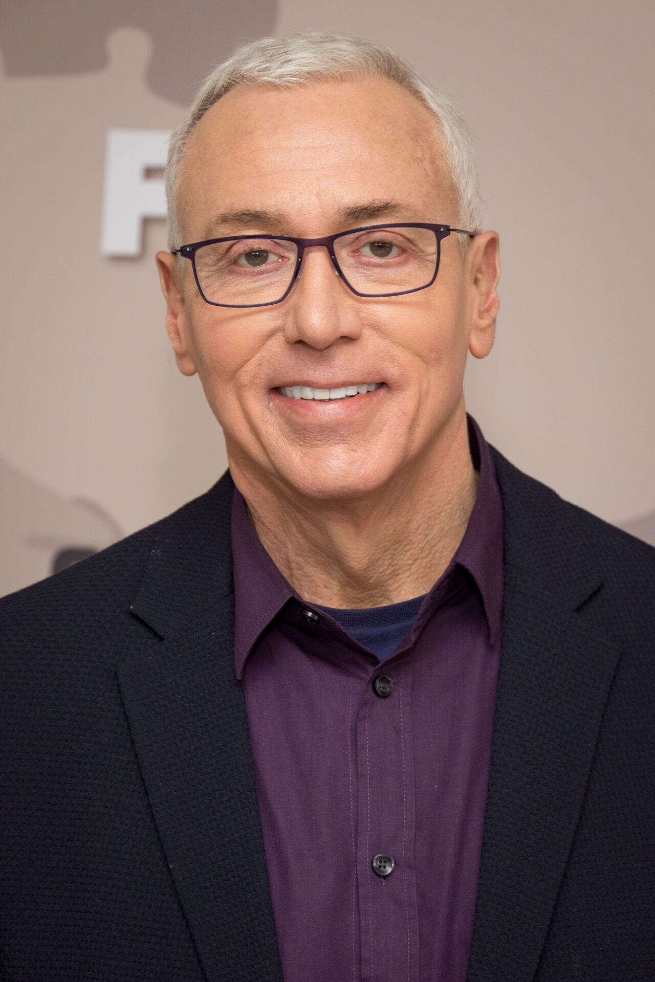 Dr. Drew smiling in a navy blazer and purple shirt, wearing rectangular-framed glasses, at a TV premiere event.
