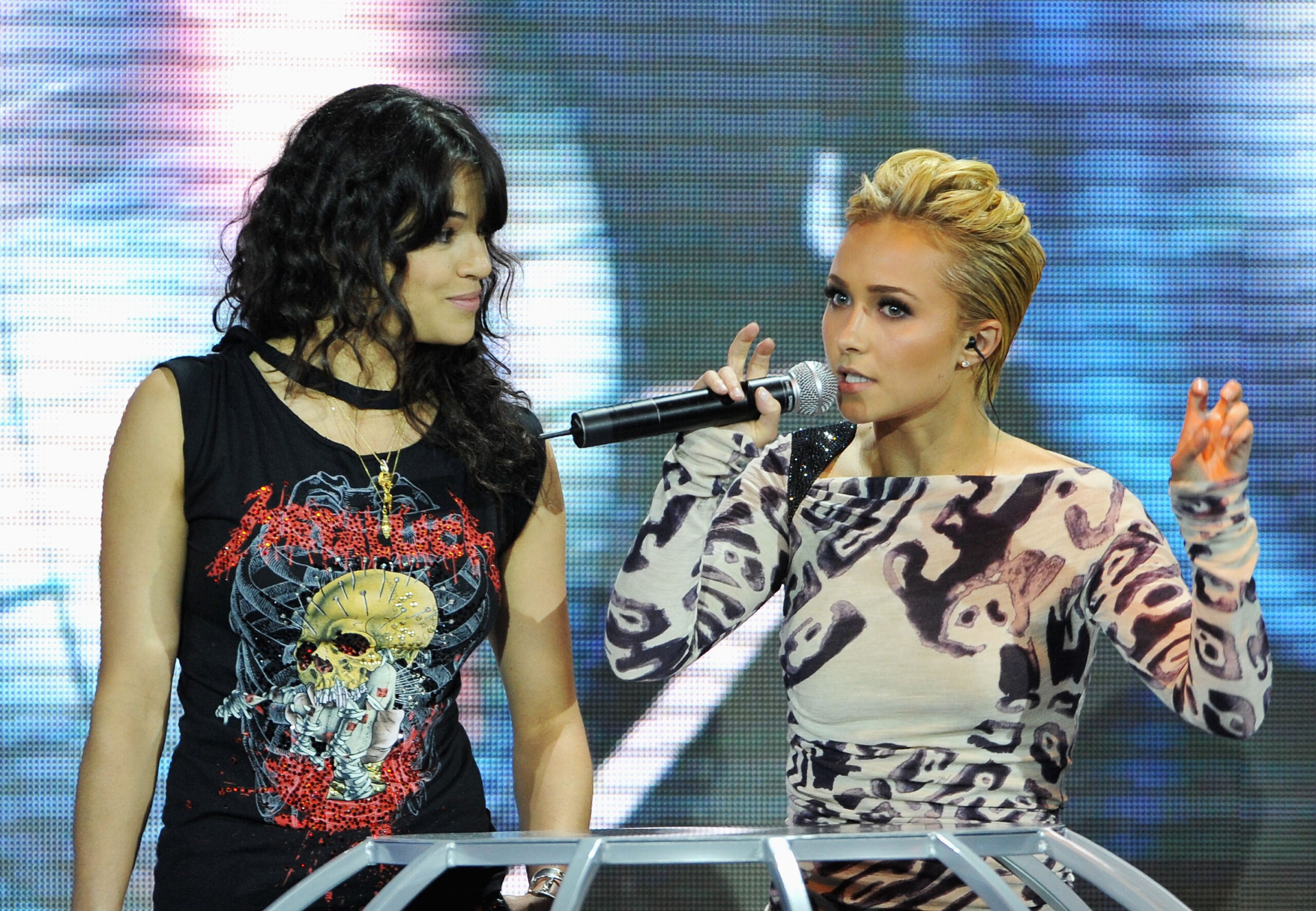 A blonde woman in a patterned dress speaks into a microphone onstage while a dark-haired woman in a black graphic tee looks on at an awards show.