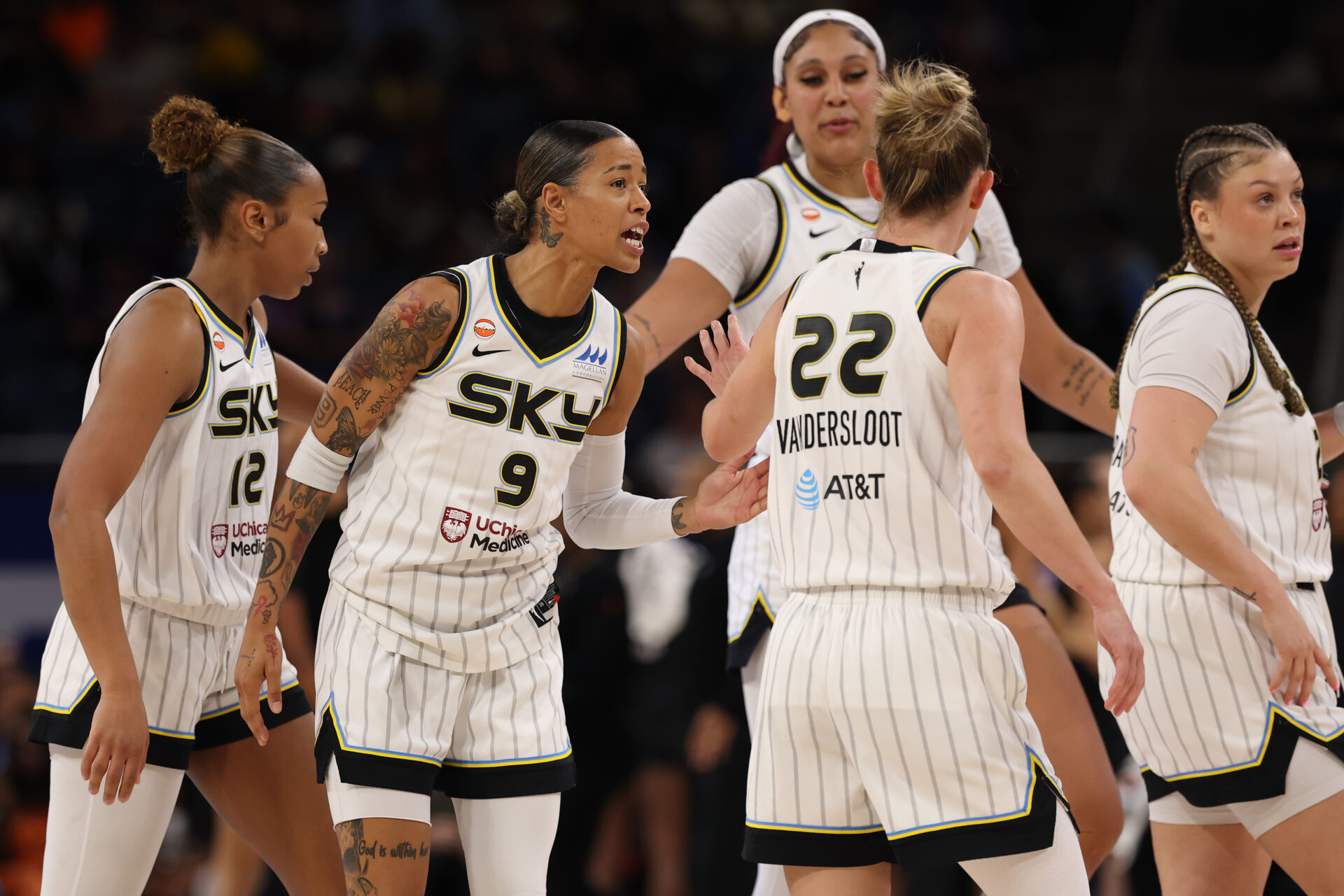 Chicago Sky players huddle on the court, with Natasha Cloud (#9) speaking to Courtney Vandersloot (#22) in white pinstripe uniforms.