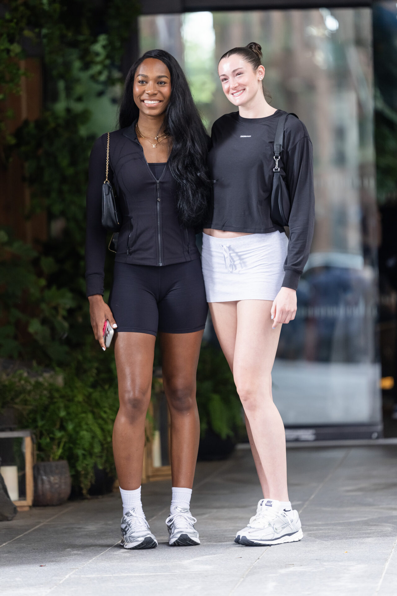 Two tall smiling women in matching black athletic outfits and white sneakers posing together on a Brooklyn sidewalk.