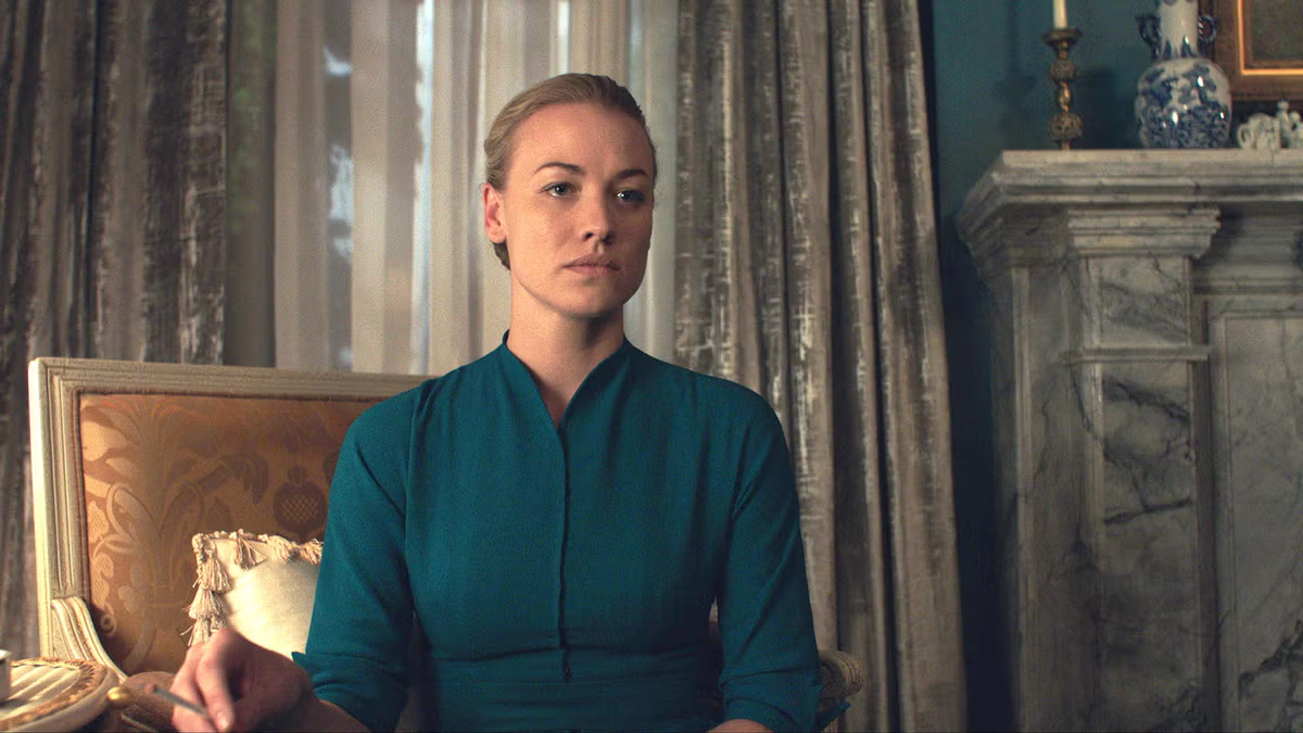Blonde woman in a teal dress seated in a gilded chair in an elegantly furnished room with marble fireplace and draped curtains