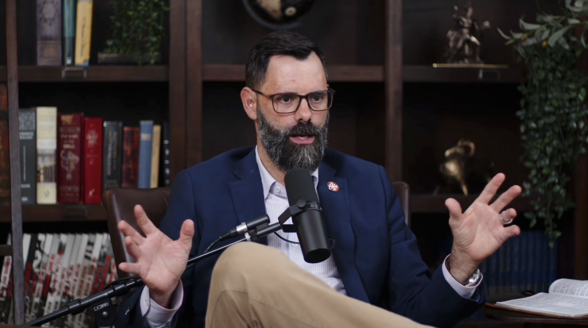 Bearded man with glasses in a navy blazer gesturing with both hands raised while speaking into a podcast microphone, bookshelves visible behind him.