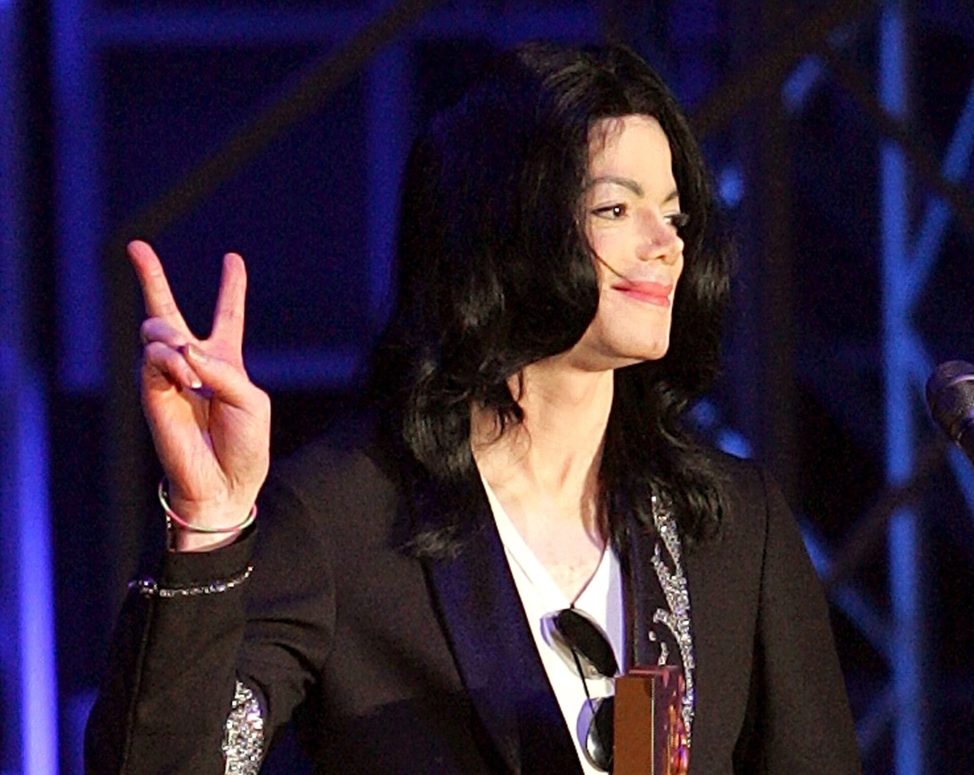 Man with long black hair in a black blazer and white top holding an award and raising a peace sign on stage.