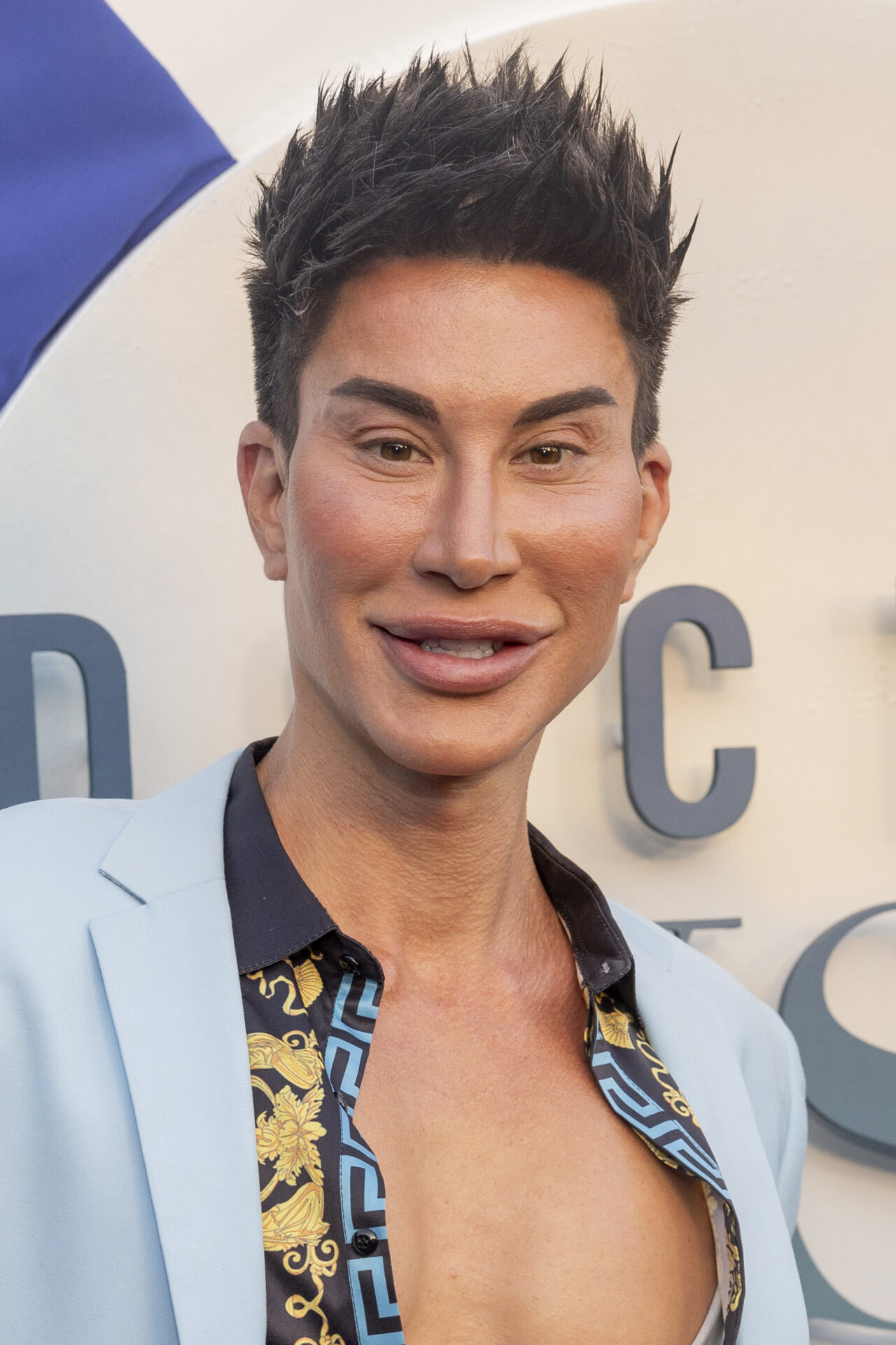 Man with spiked dark hair wearing a light blue blazer over an open printed shirt, smiling at a premiere event.