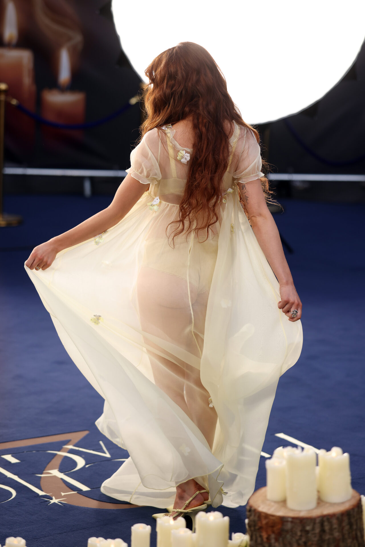 Woman with long auburn hair wearing a sheer cream floral maxi dress on a blue carpet surrounded by pillar candles, seen from behind.