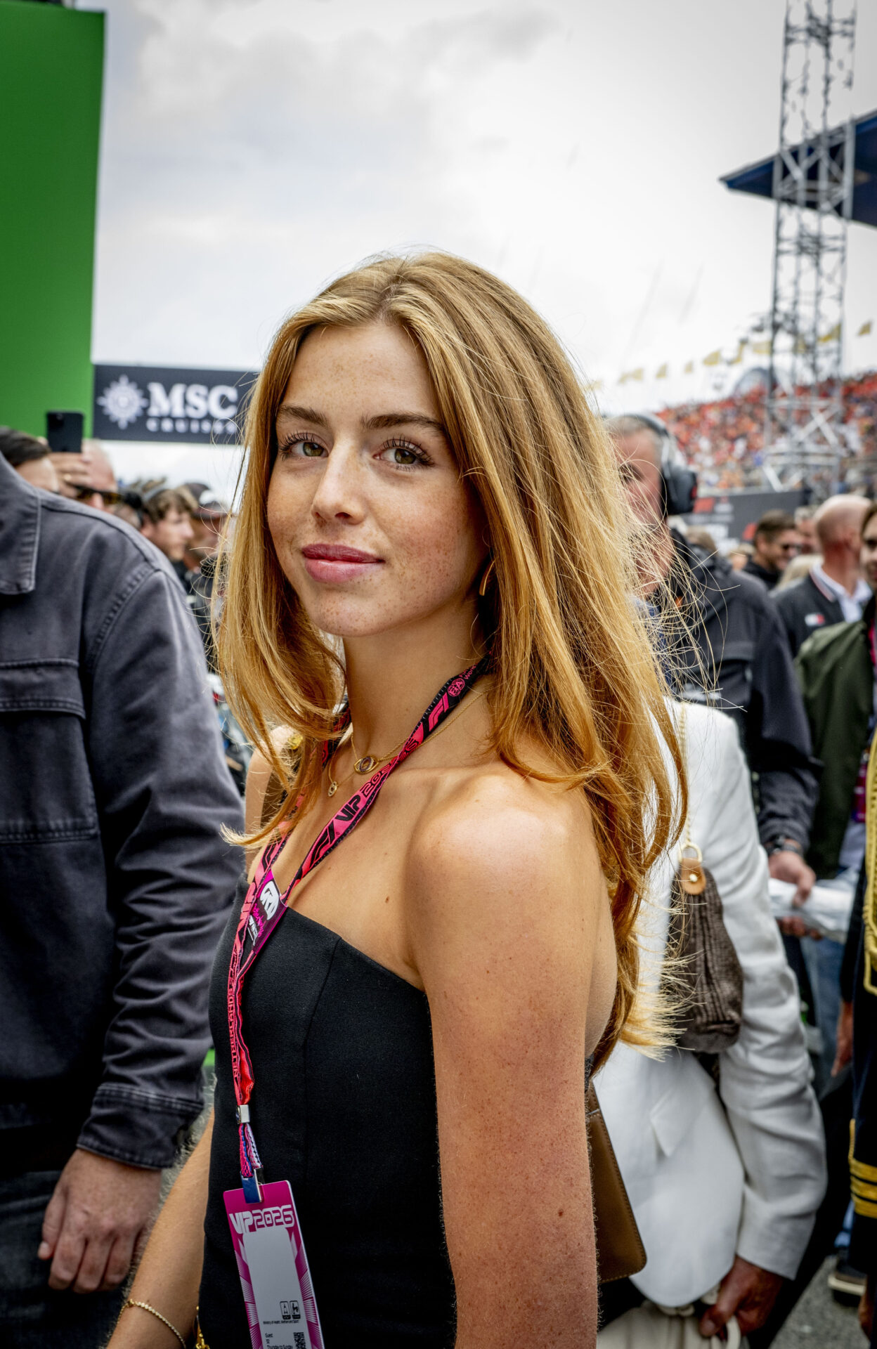 Princess Alexia in a black strapless dress with an F1 VIP lanyard, looking back over her shoulder on a busy F1 grid at Zandvoort.