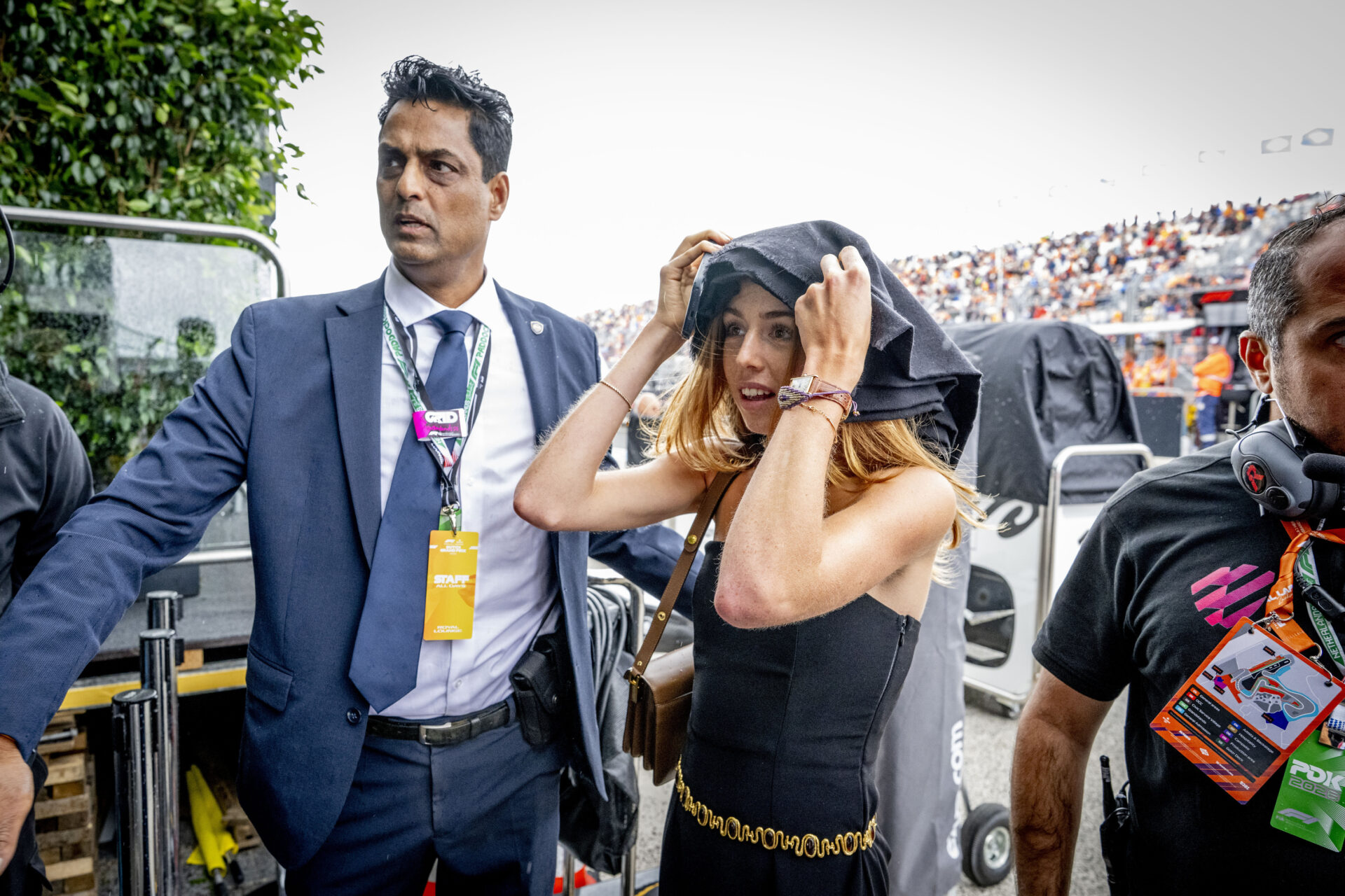 A young woman in a black strapless dress pulling a jacket over her head in the rain on the F1 grid, flanked by staff members at Zandvoort.