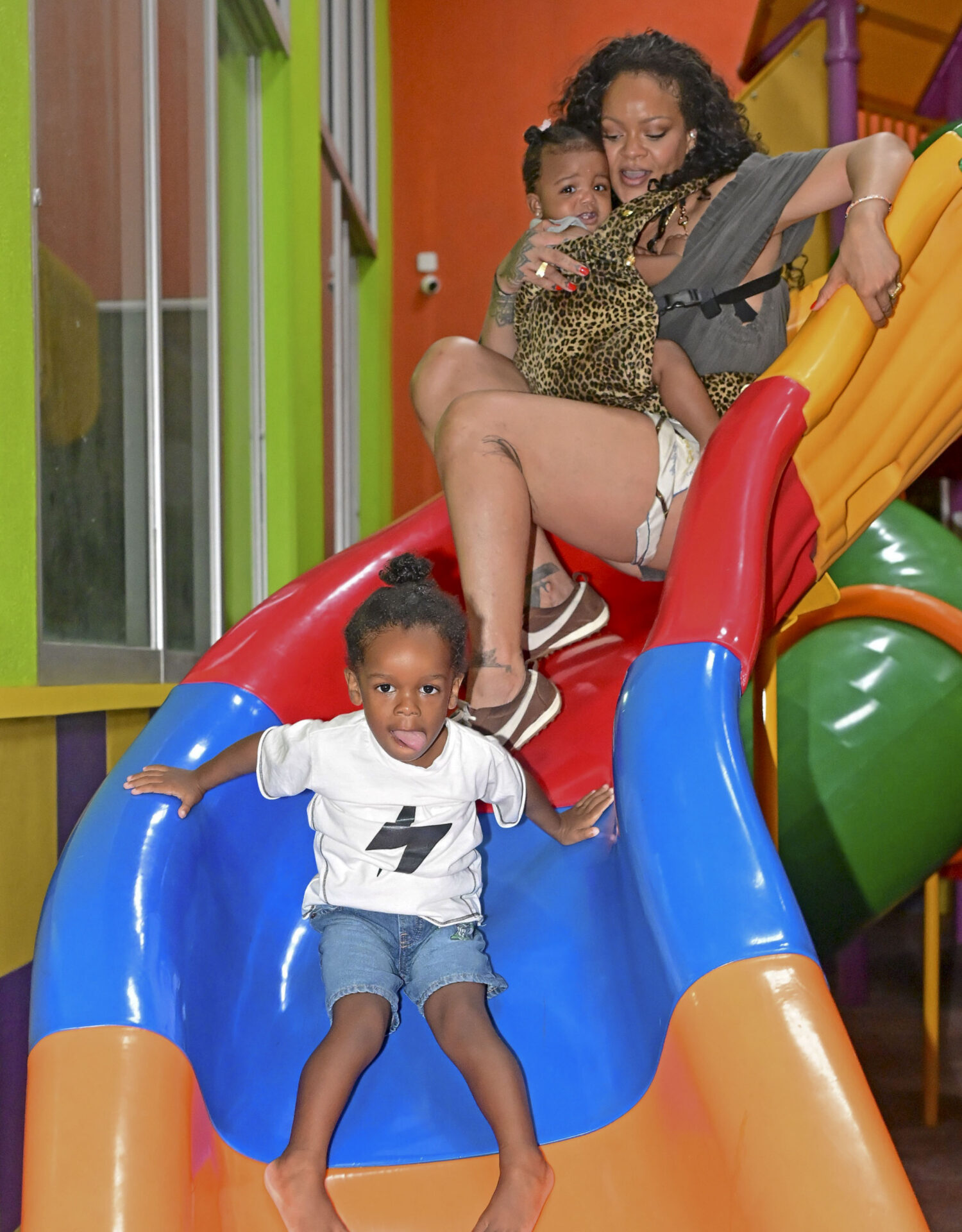 A woman holding a baby in a leopard-print carrier slides down a red and blue indoor playground slide while a young boy in a white shirt goes down in front of her.