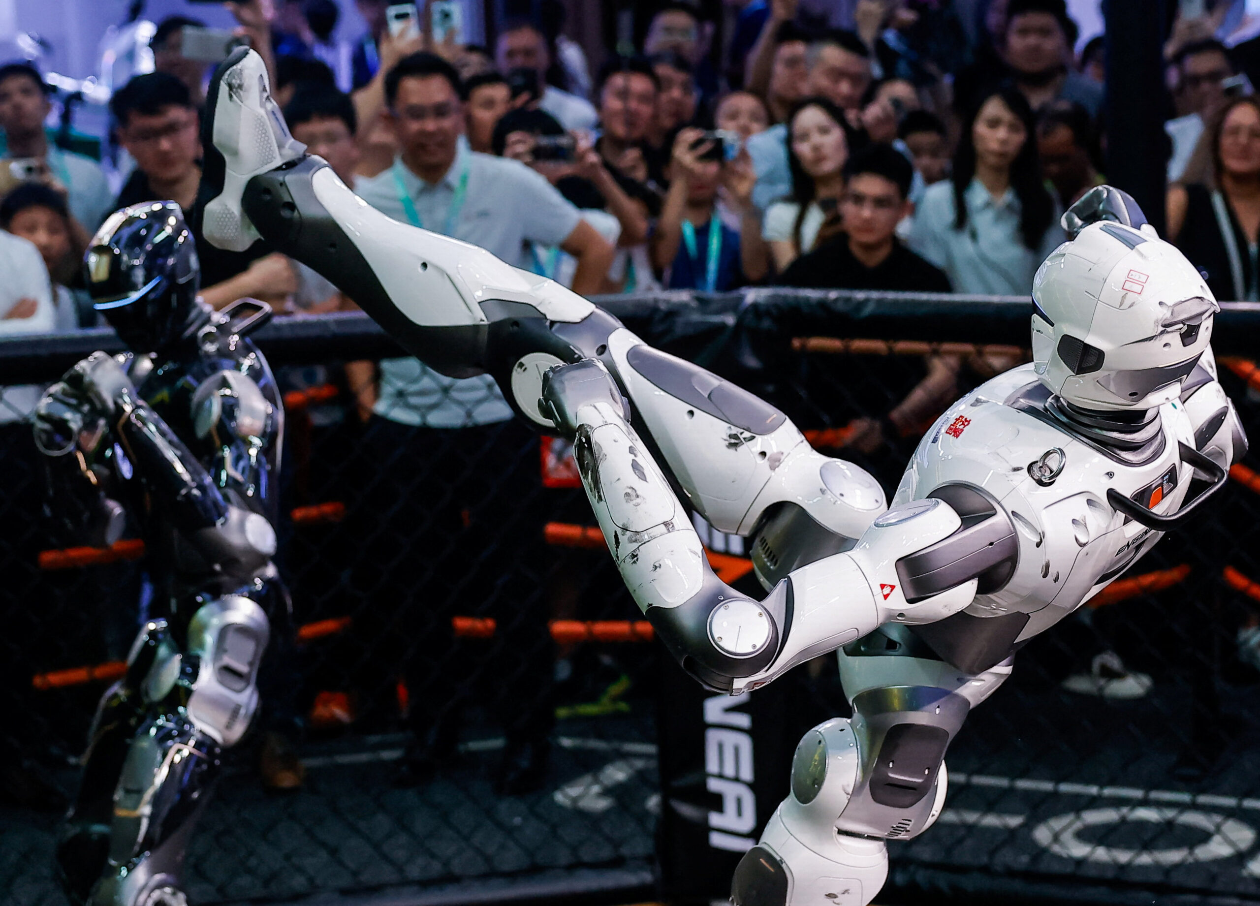 A white humanoid robot with one leg raised in a high kick faces a darker robot inside an octagon-style cage as spectators watch.