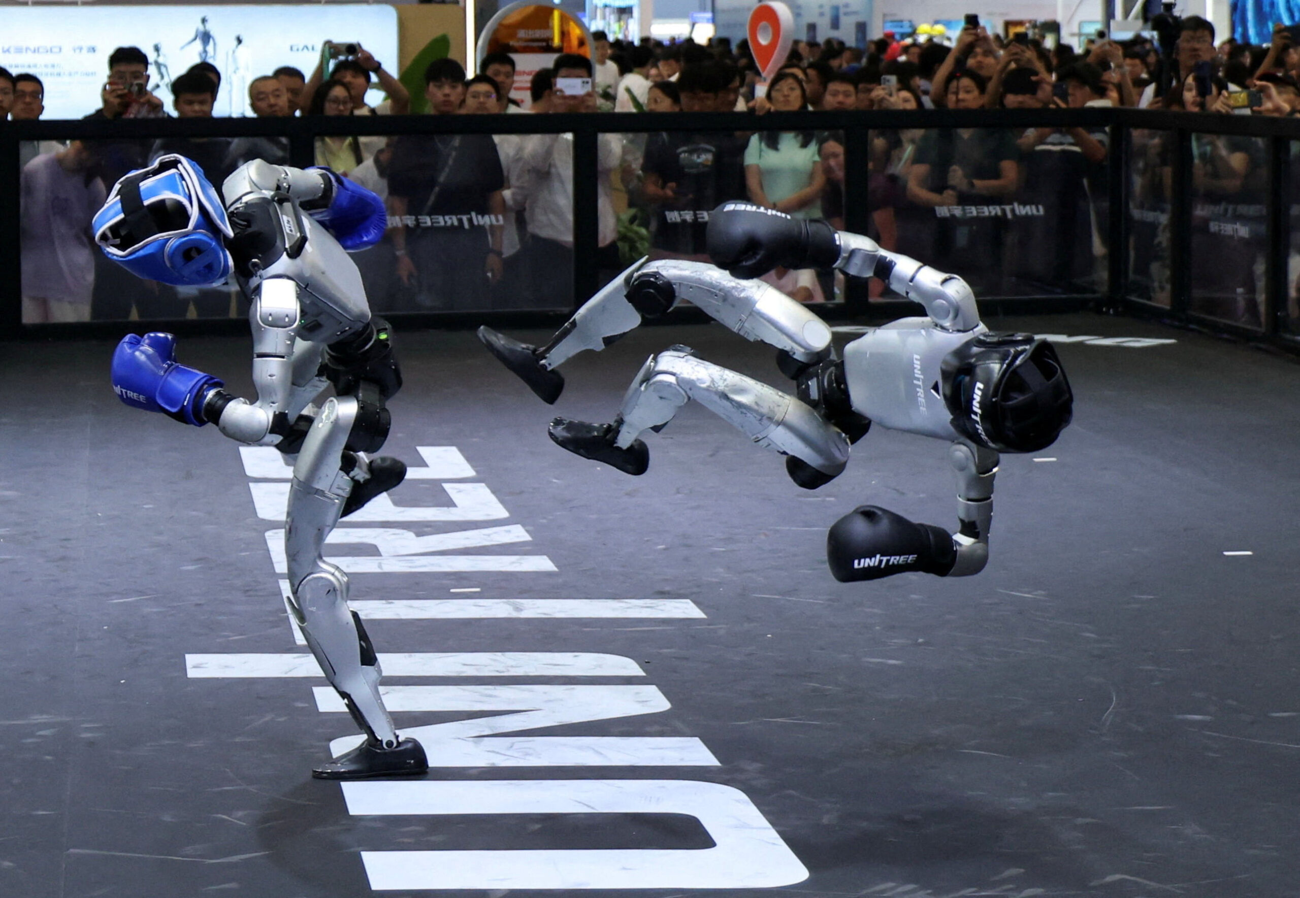 A humanoid robot in blue boxing gloves kicks a second robot, which is sent flying sideways in mid-air, before a packed crowd.