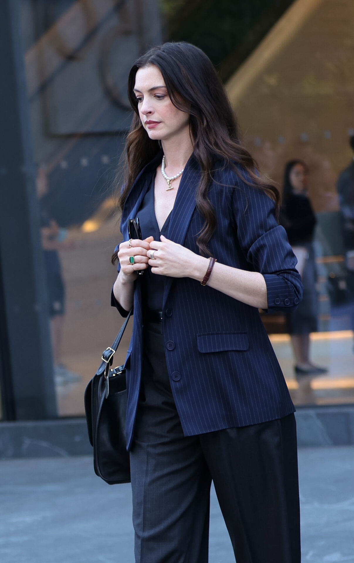 Anne Hathaway in a navy pinstriped blazer, dark wide-leg trousers, and pearl necklace, holding sunglasses and a black handbag on a city street.