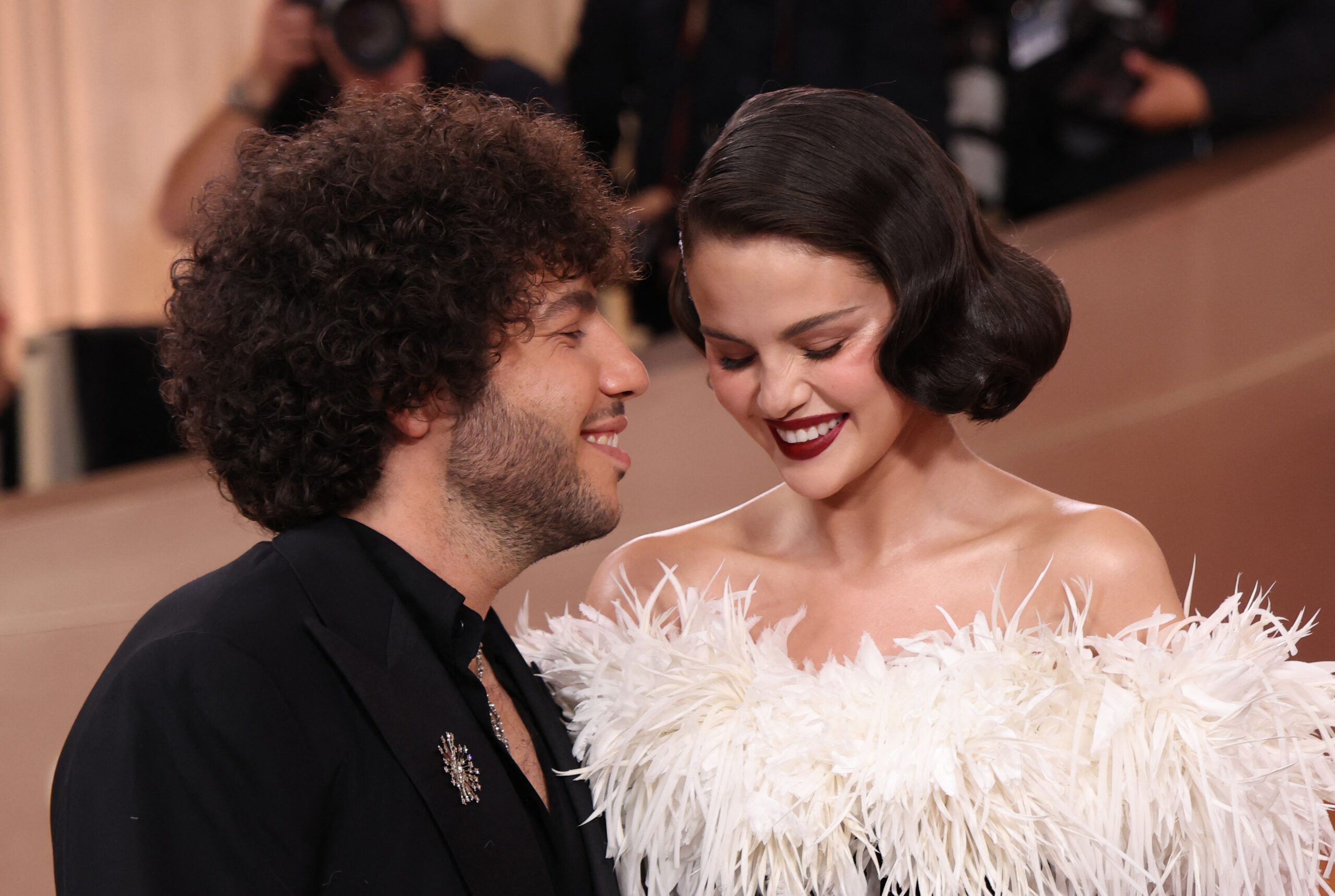 Benny Blanco and Selena Gomez smiling at each other at the Golden Globes red carpet, she in a white feathered gown.