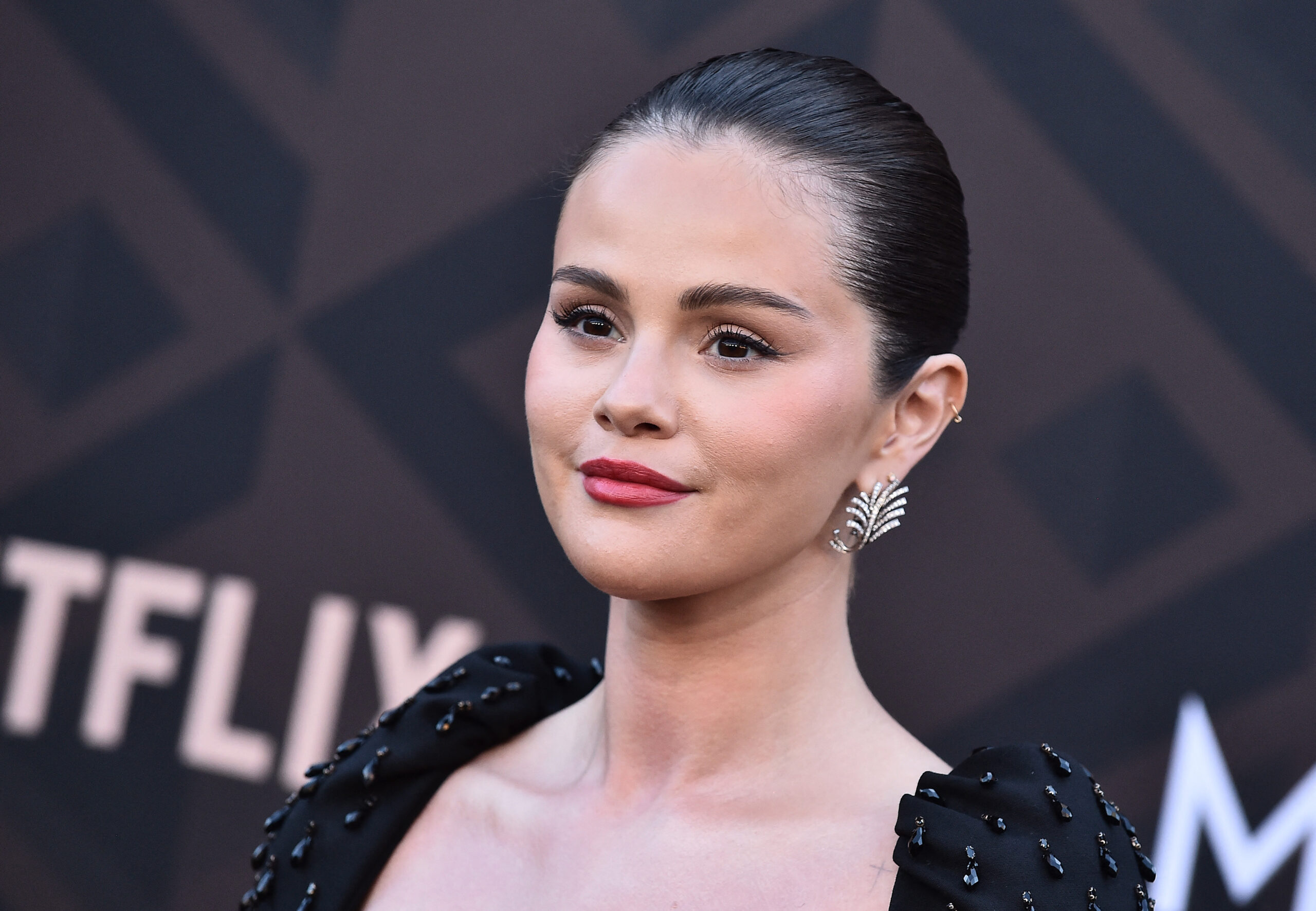 Selena Gomez in a black beaded off-shoulder dress with slicked-back hair and red lips at a Netflix premiere event.