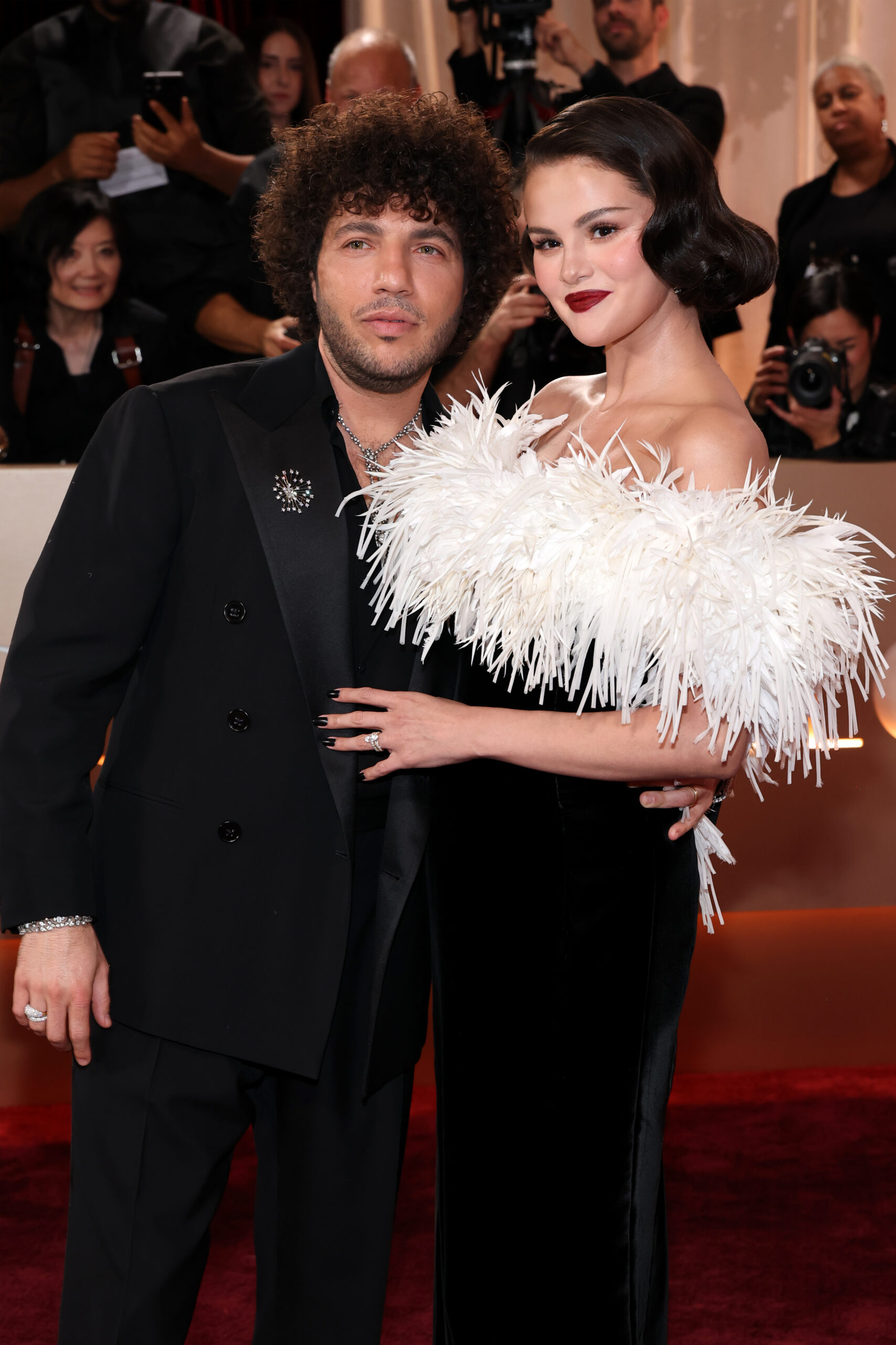 A man in a black suit and a woman in a black velvet dress with a white feathered top pose arm-in-arm on a red carpet.