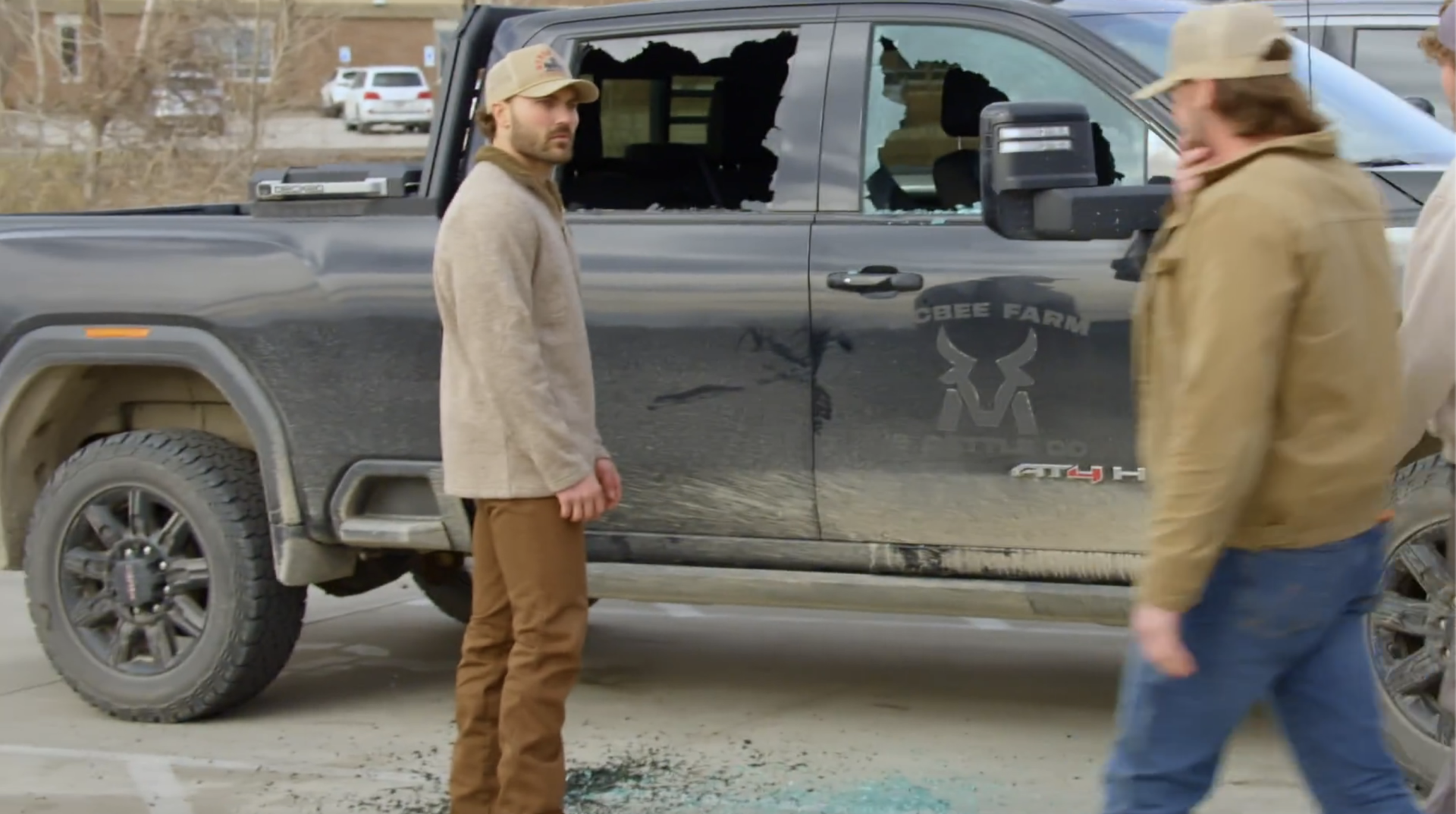 Man in beige pullover and tan cap standing next to a damaged gray GMC AT4 farm truck with two broken cab windows and glass on the ground.