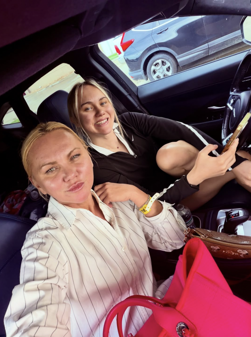 Two blonde women smiling inside a car; one takes a selfie while the other holds a phone, with a pink bag in the foreground.
