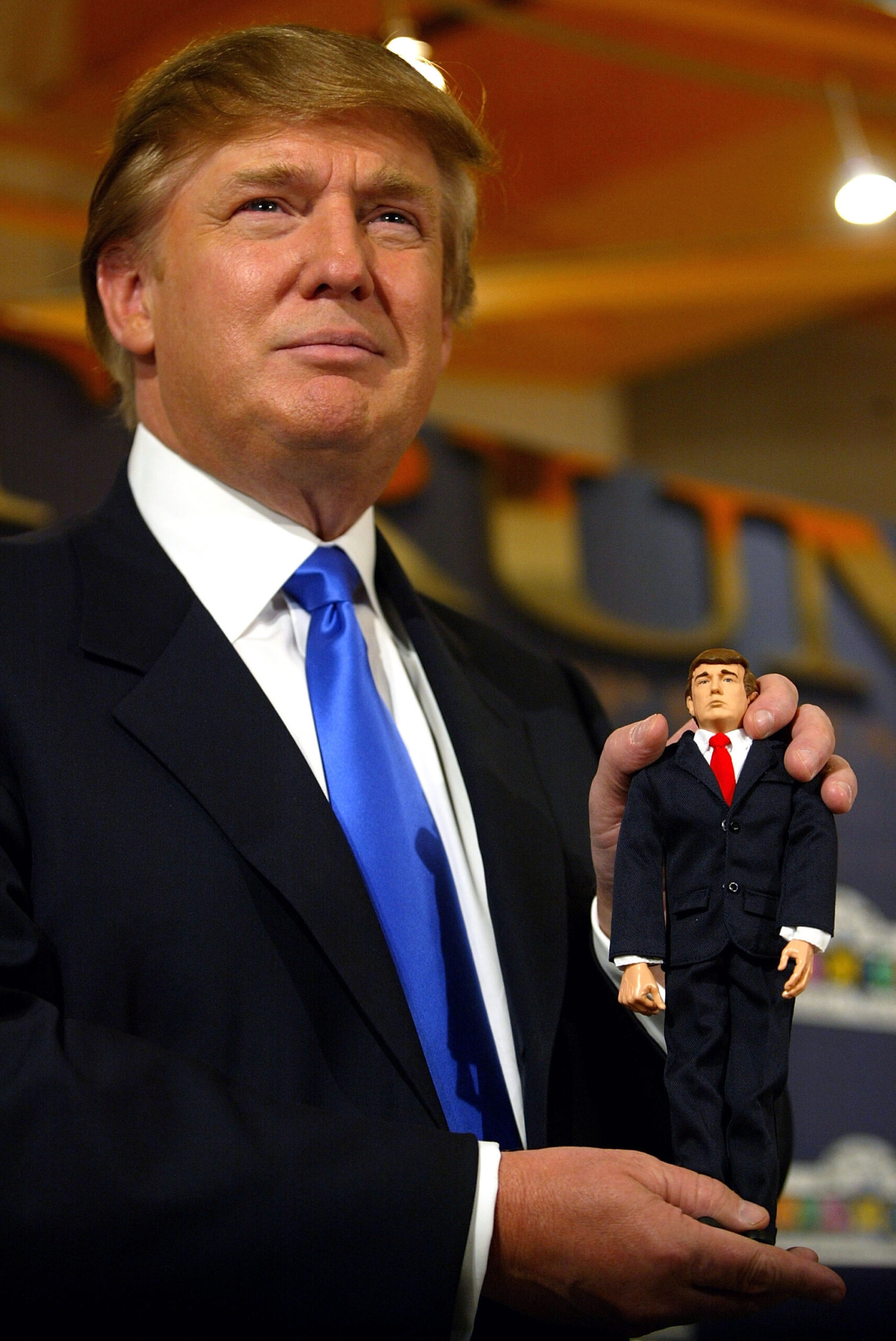 Man in dark suit and blue tie holds a small action figure dressed in a suit at a toy store event.