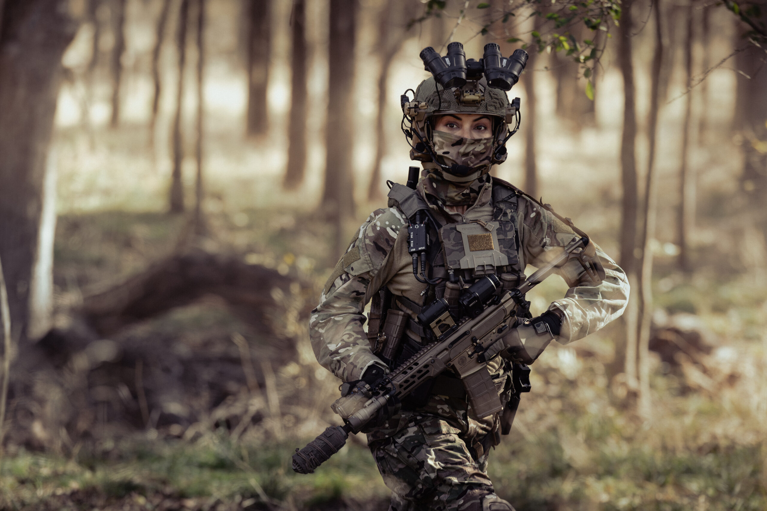 Female soldier in full camouflage gear with night-vision helmet, tactical vest, and rifle standing in a forest.