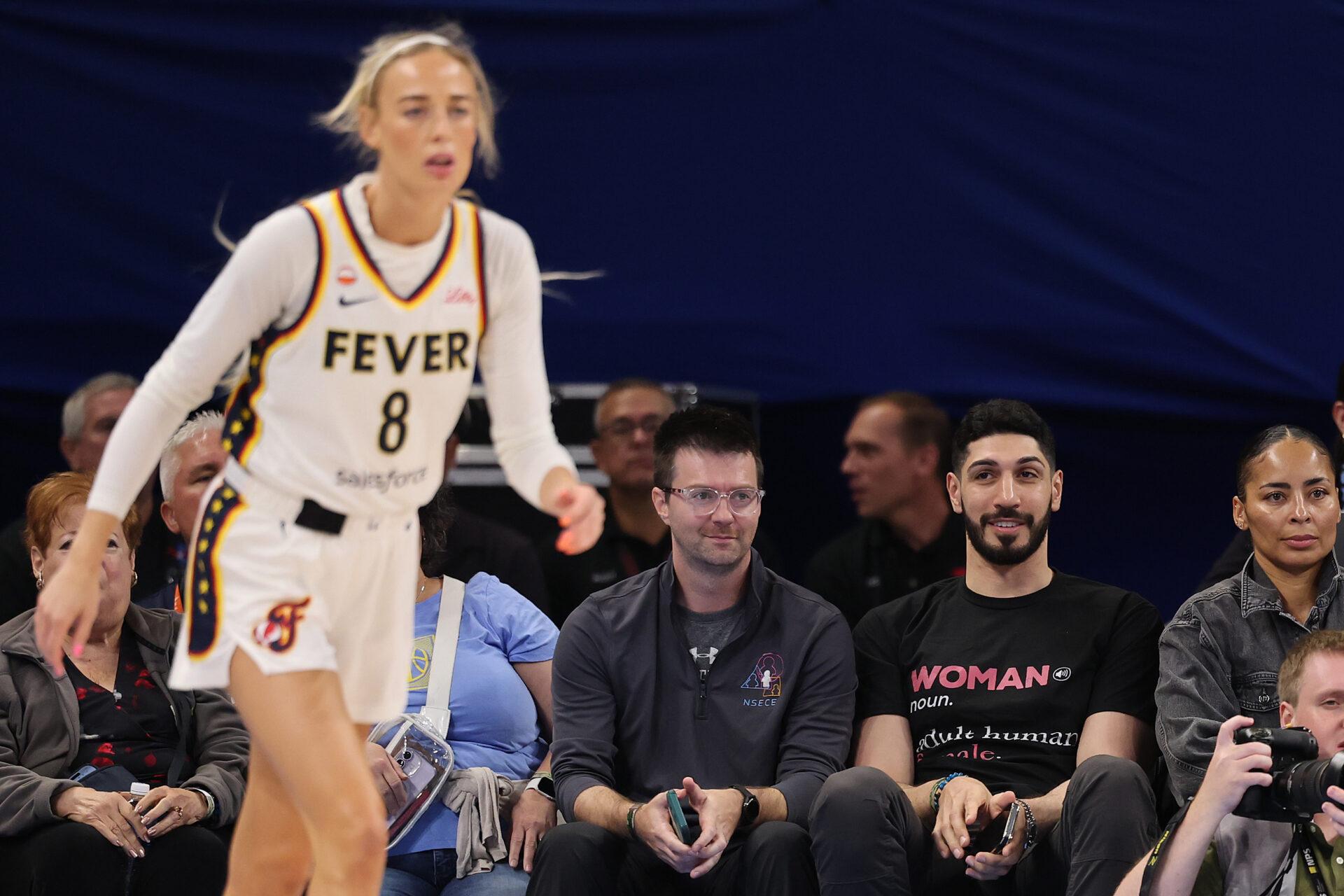Indiana Fever player Sophie Cunningham number 8 runs on court in the foreground as Enes Kanter Freedom watches from a courtside seat.
