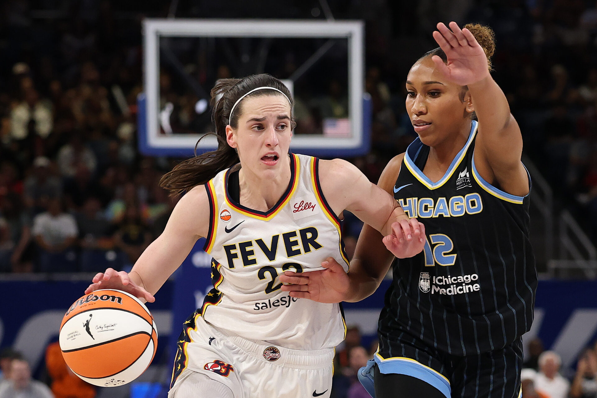 Caitlin Clark in a Fever jersey dribbles toward the basket while Chicago Sky's Sydney Taylor #12 defends with an outstretched arm.