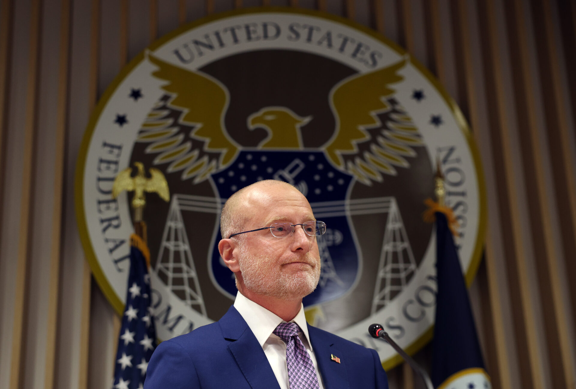 Bald man with glasses and blue suit speaking at a podium in front of the Federal Communications Commission seal and American flags.
