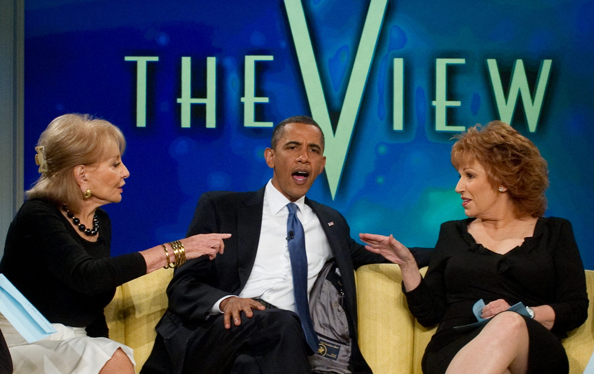A man in a suit and blue tie gestures while seated between two women on The View set, with the show's logo visible behind them.