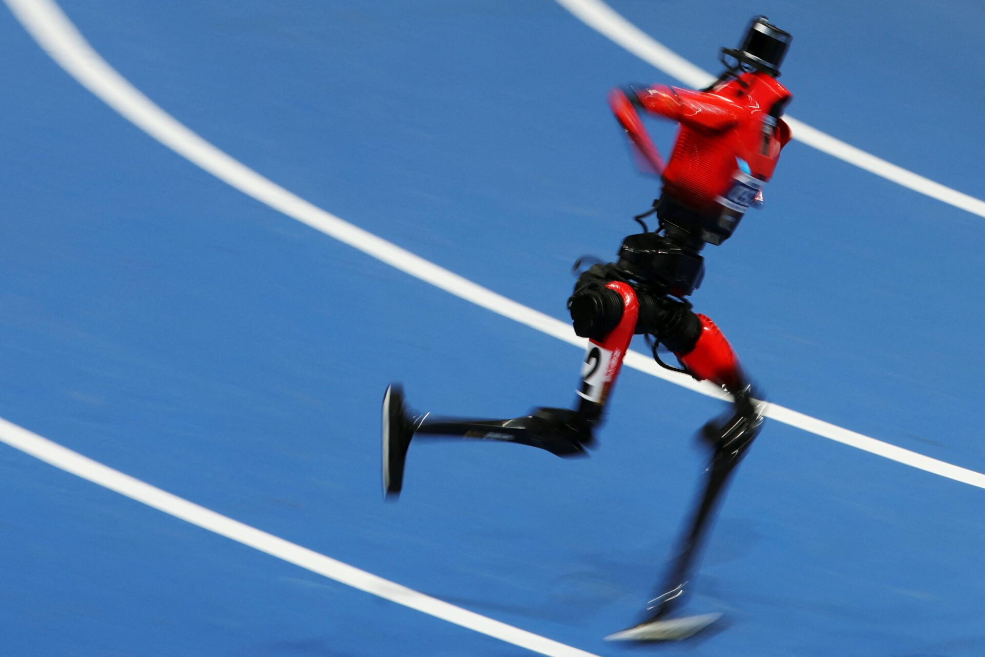 A red humanoid robot with blade-like feet running at speed on a blue athletics track, heavily motion-blurred, wearing race number 2.