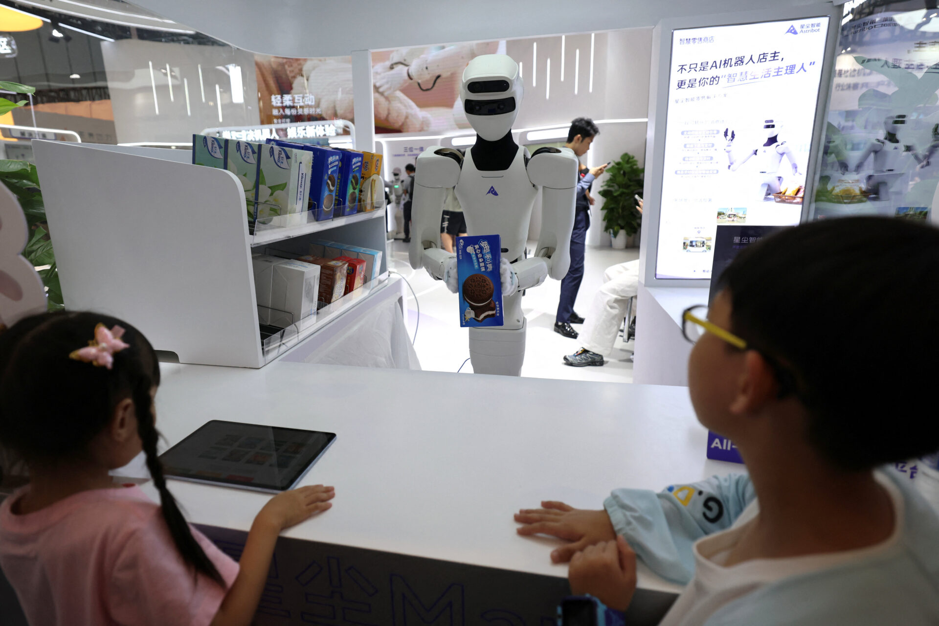 Two children at a counter look up at a white humanoid Astribot robot holding a cookie package in a mock retail shop at a Beijing tech conference.
