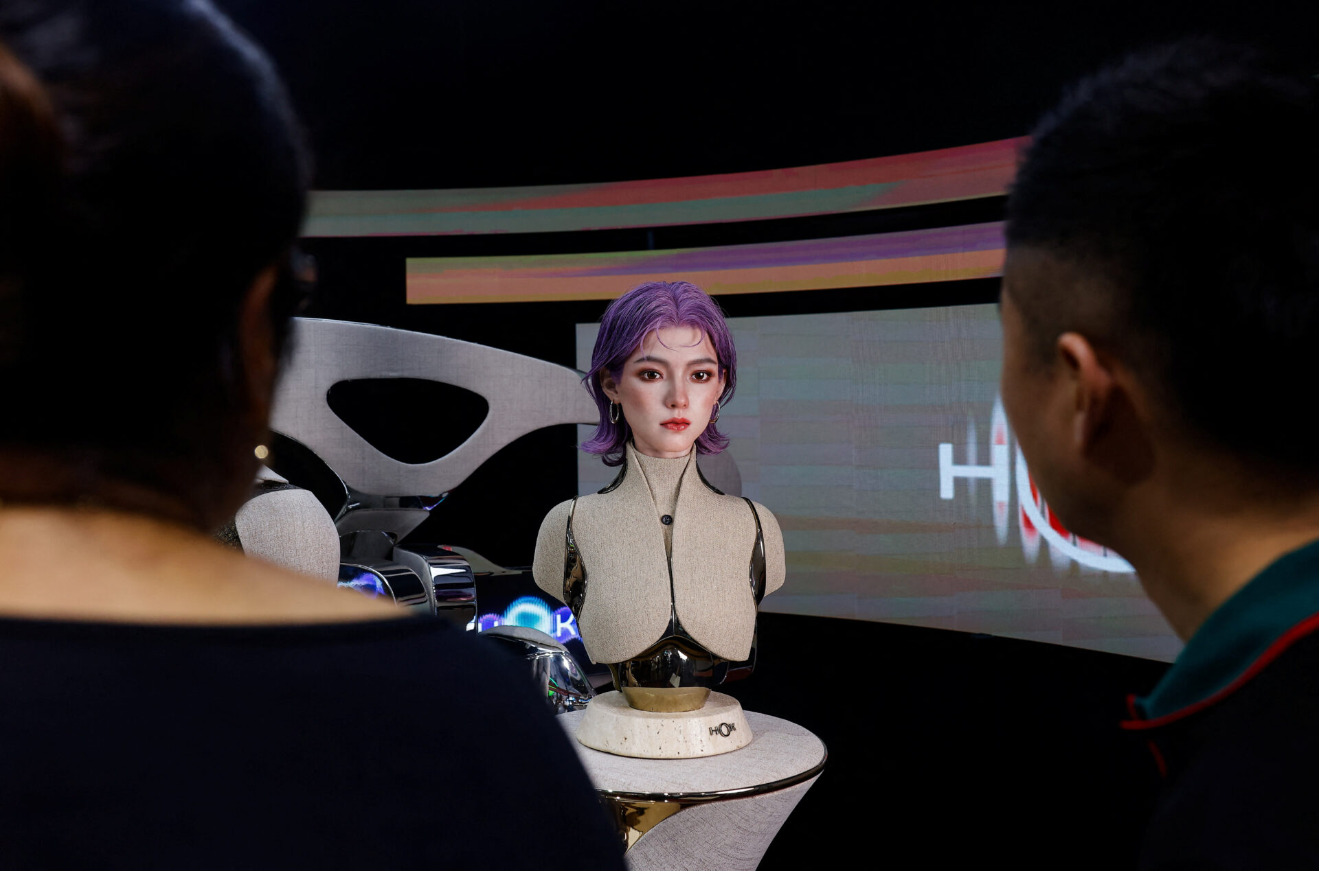 Two people seen from behind looking at a purple-haired humanoid robot mounted on a pedestal, with colorful display screens in the background.