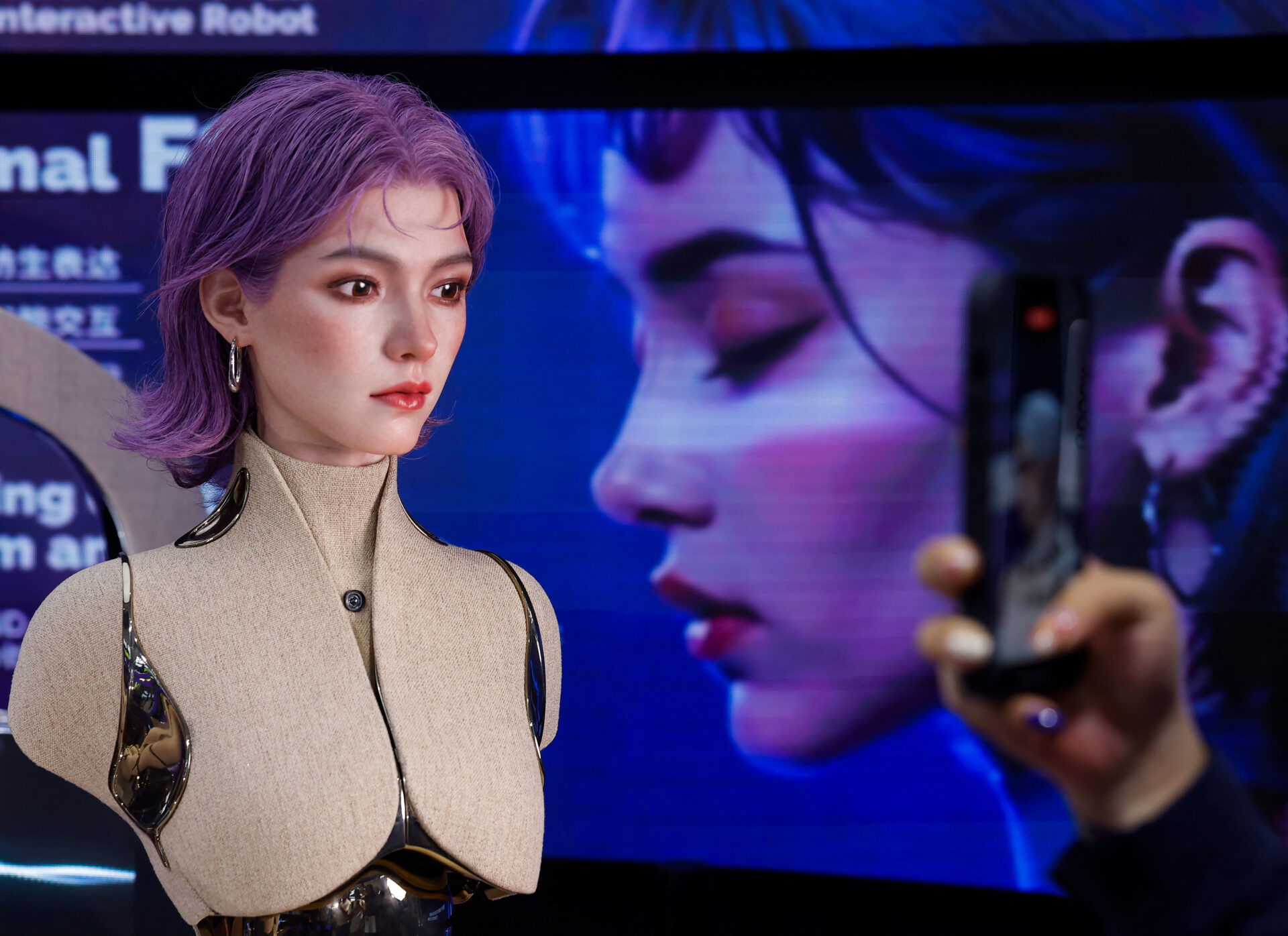 Close-up of a lifelike humanoid robot with purple hair and metallic body panels, with a hand holding a smartphone filming it in the foreground.