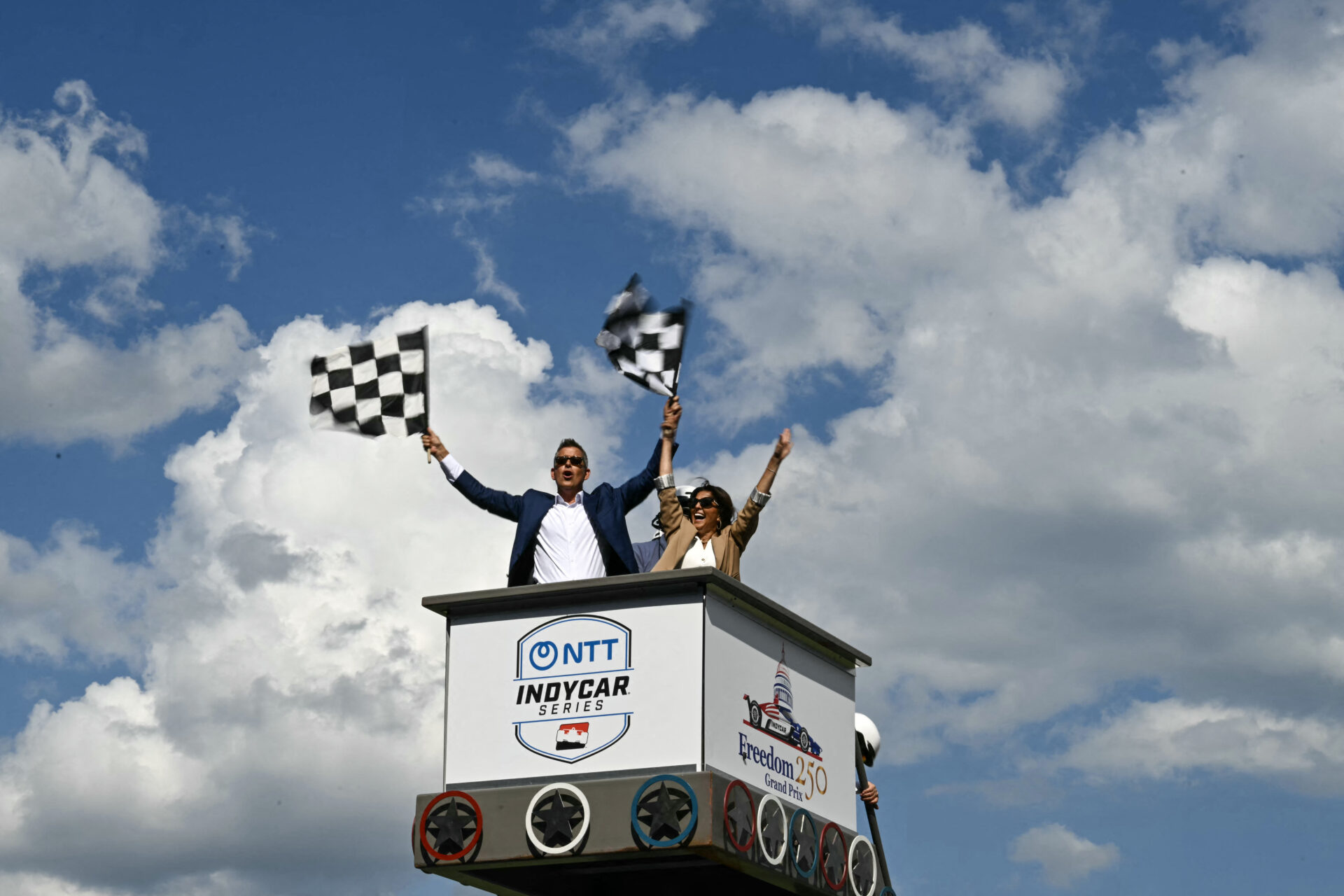 Sean Duffy and Rachel Campos-Duffy wave checkered flags from an elevated NTT IndyCar Series starter's stand under a partly cloudy sky.
