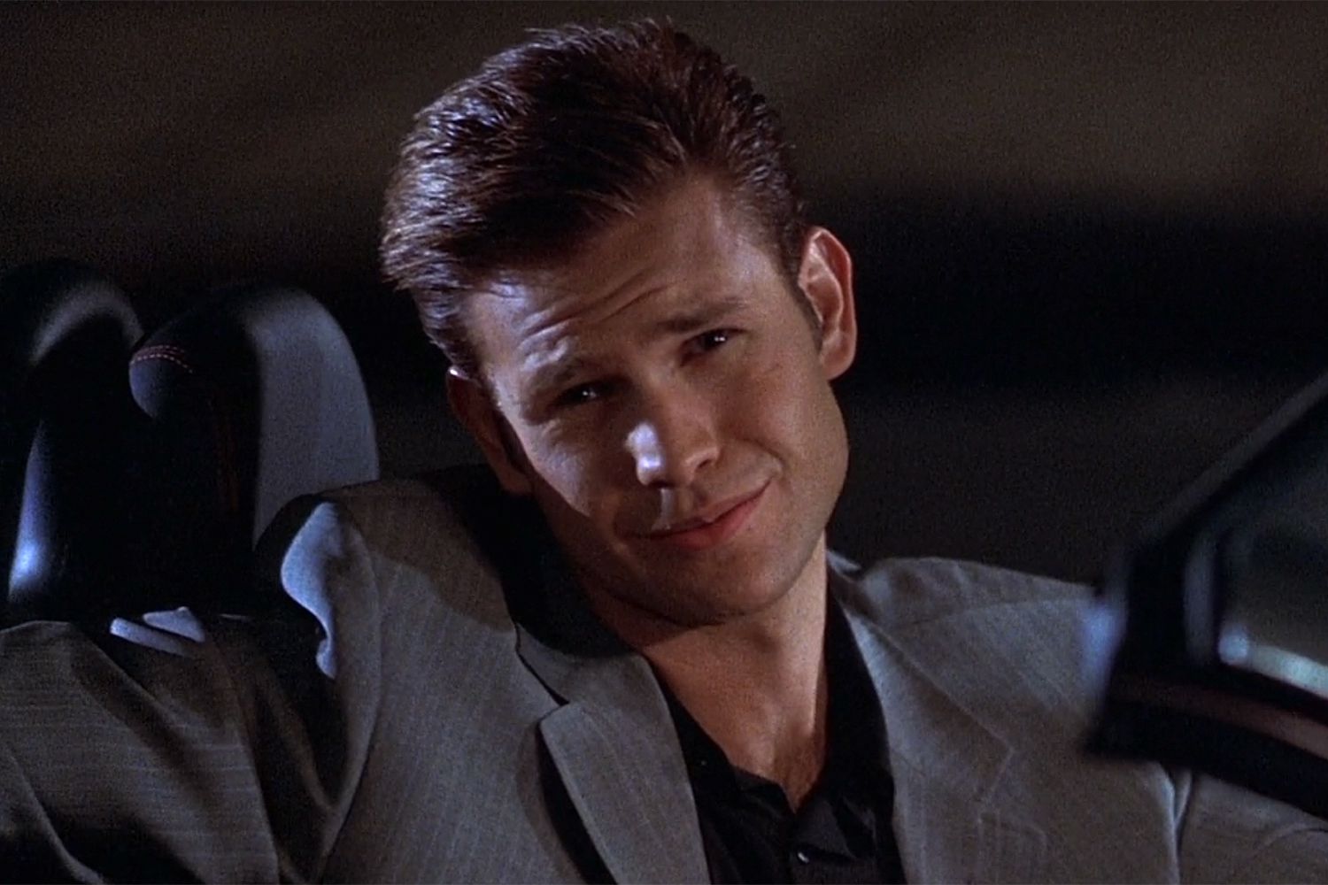Young man with slicked-back brown hair wearing a grey blazer smirking in the back seat of a car.