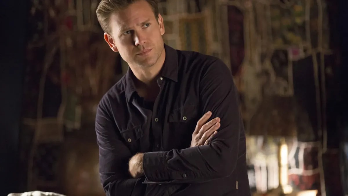 Blond man with arms crossed wearing a dark button-up shirt, standing in front of a wall covered in papers and writing.