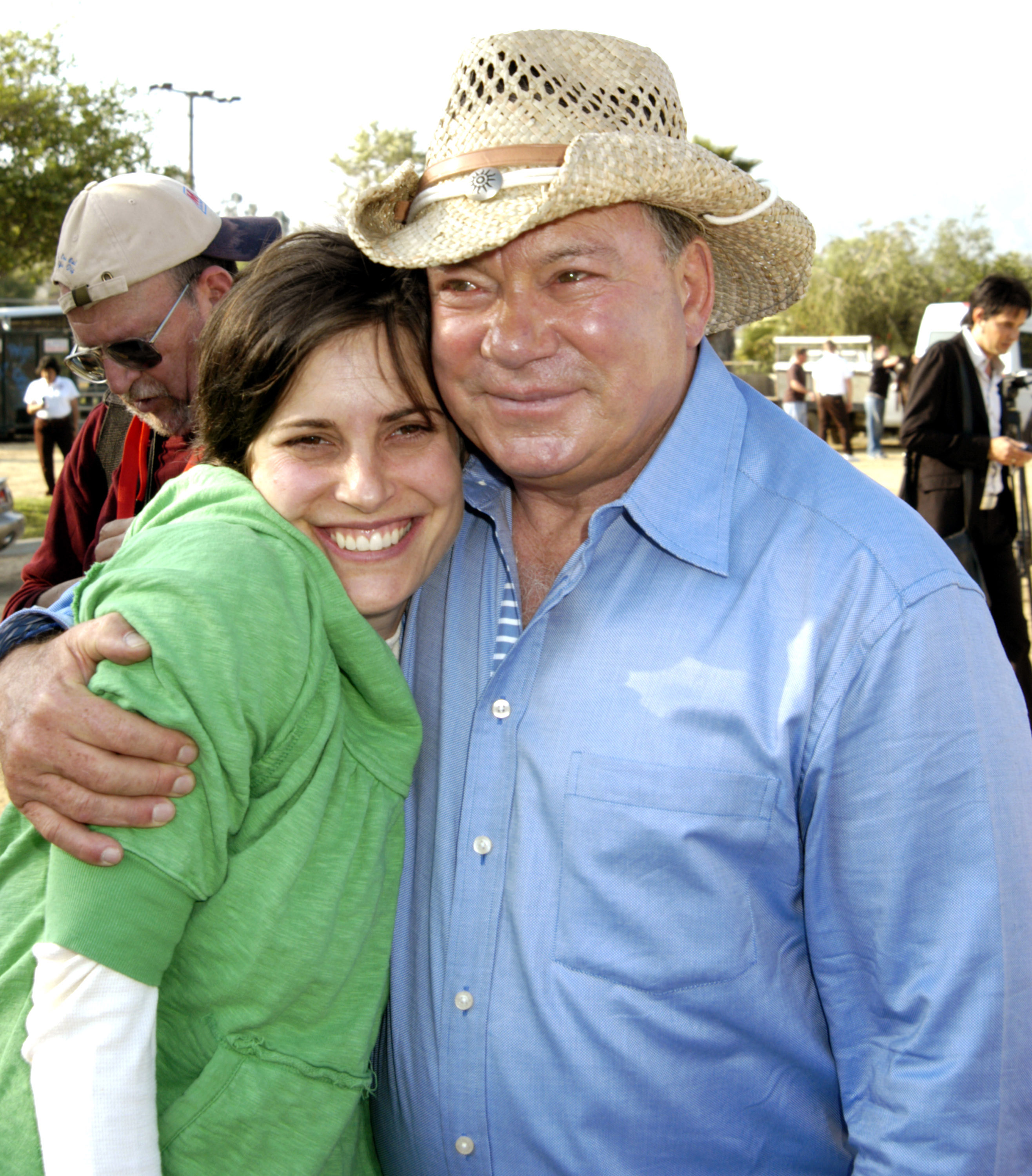 Melanie Shatner in a green hoodie smiles as William Shatner, wearing a straw cowboy hat and blue shirt, hugs her at an outdoor equestrian event.