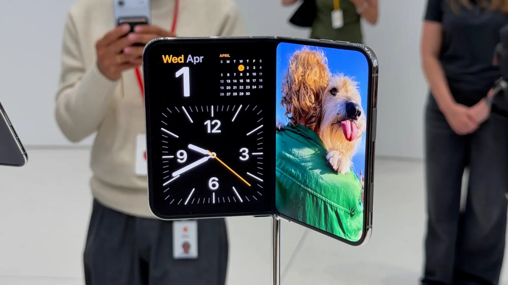 An unfolded iPhone Duo sits partially folded on a display stand, showing StandBy mode with a calendar and clock on one half and a photo of a dog on the other.