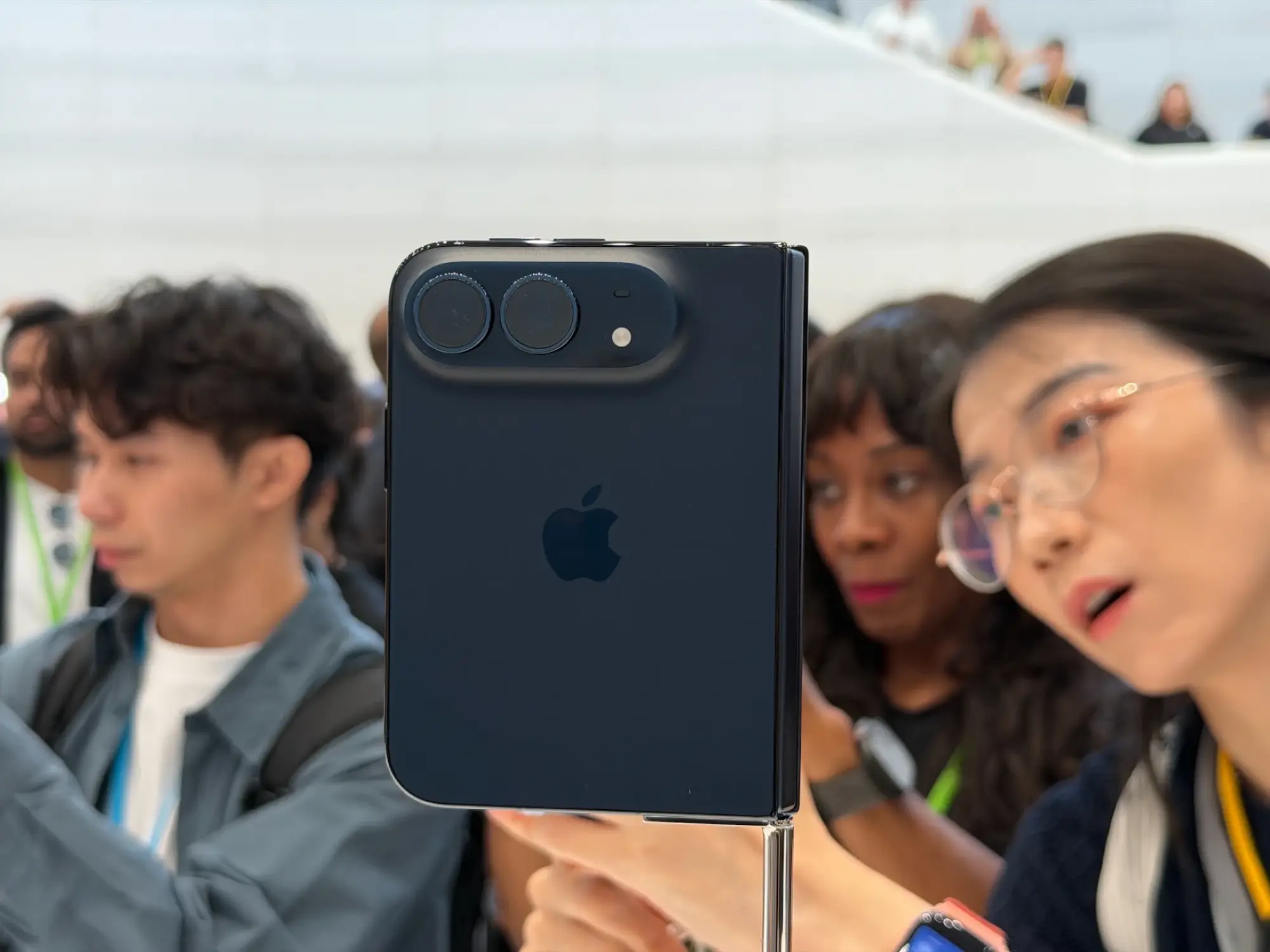 Rear view of a folded iPhone Duo in Night Sky on a display stand, showing its dual cameras and side hinge as attendees look on.